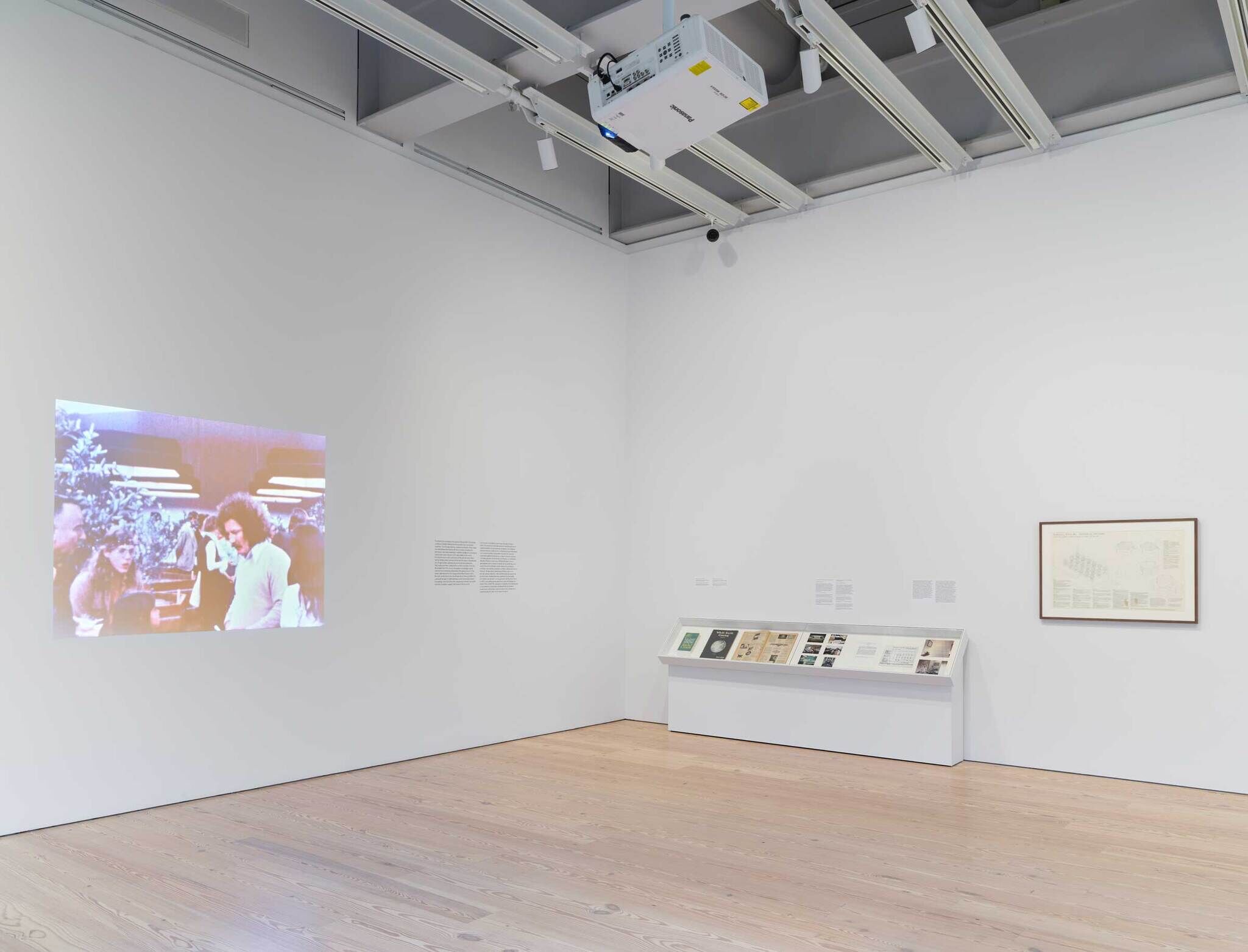 A gallery room with a video projection on the left wall, a display case with documents in the center, and a framed artwork on the right wall.