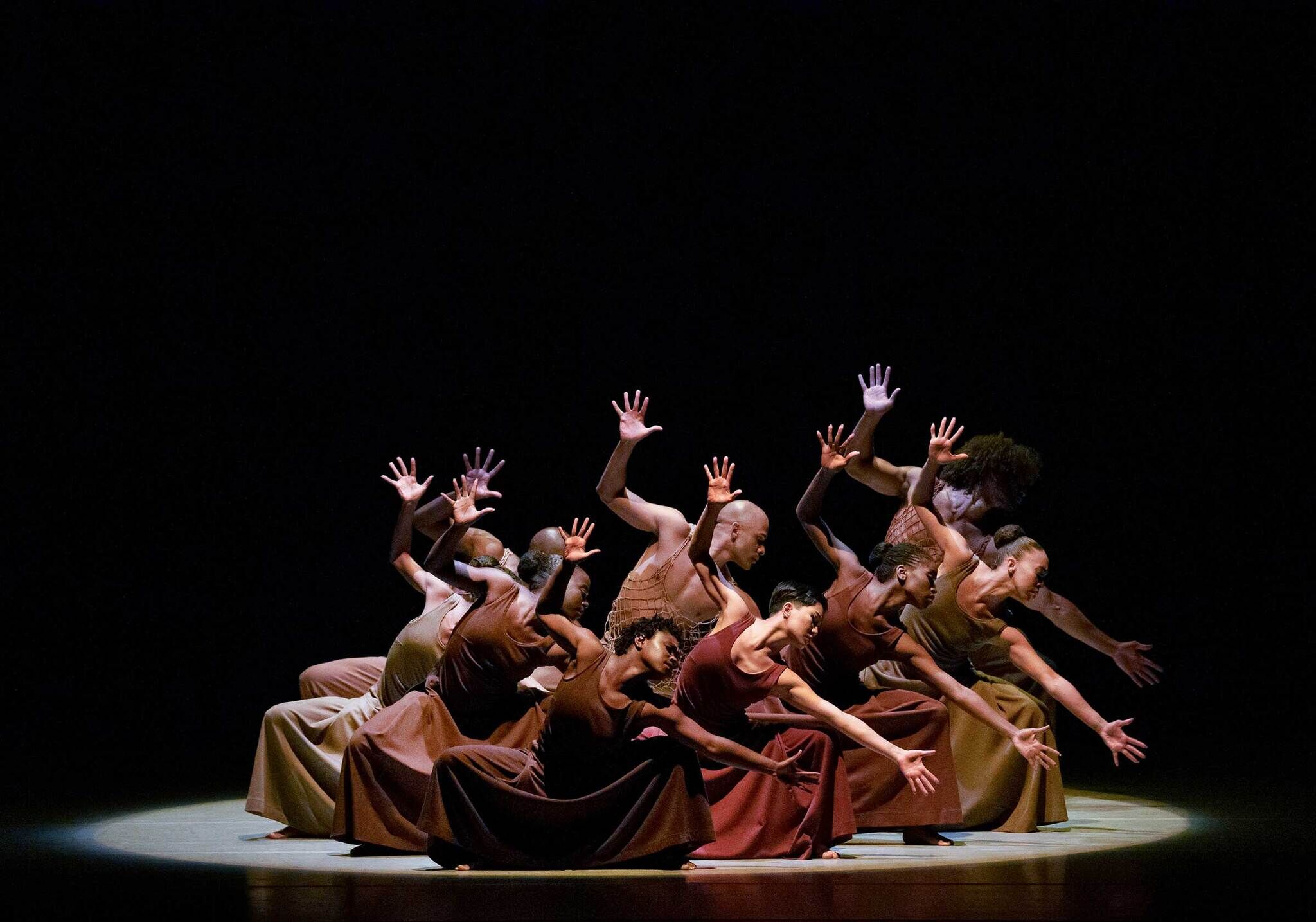 A group of dancers in brown costumes perform on stage, illuminated by a spotlight, with their arms raised and bodies gracefully bent.