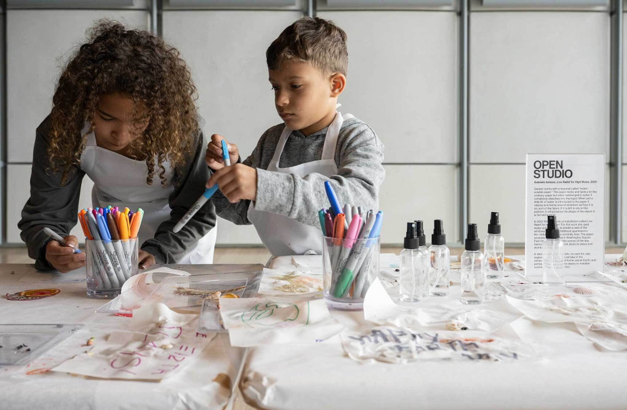 Two children in aprons are drawing with markers at a table filled with art supplies, including spray bottles and a sign that reads "Open Studio."