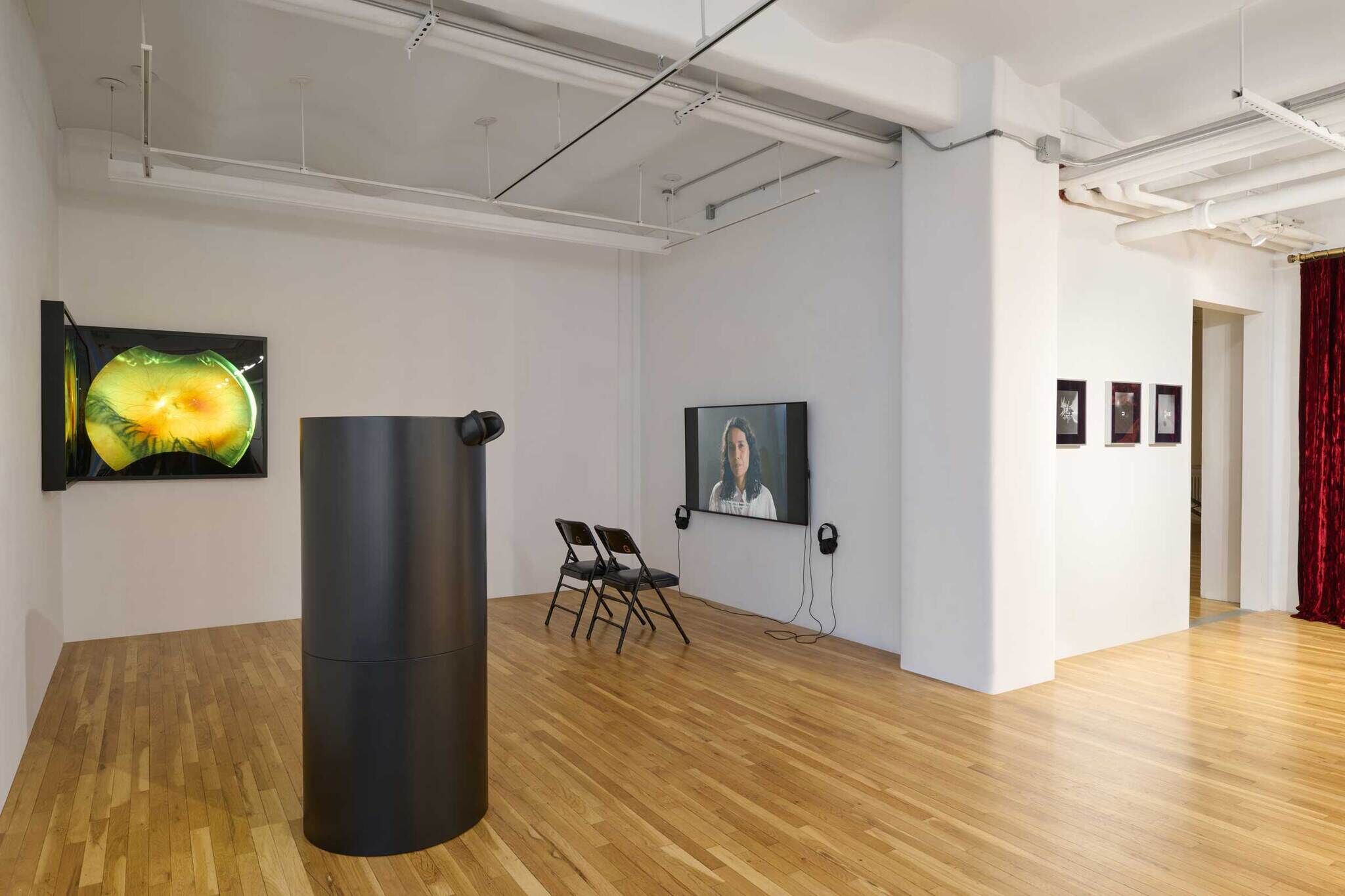 Modern art gallery with wooden floors, featuring two screens, a cylindrical black sculpture, and framed artworks on the wall.