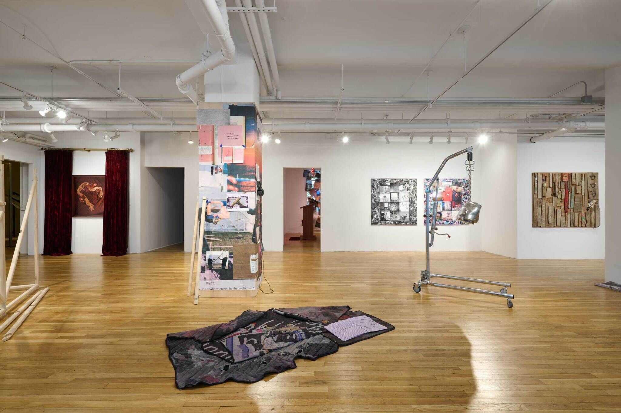 Art gallery with various installations, including a hanging sculpture, wall art, and a fabric piece on the wooden floor. Red curtains in the background.