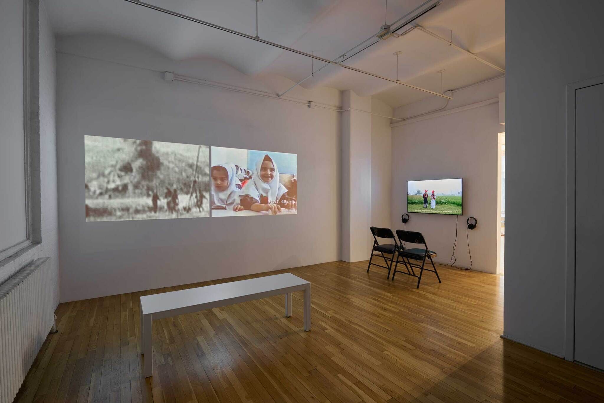 A minimalist gallery room with two video projections on one wall and a TV screen on another, showing various scenes. Two chairs and a bench are present.