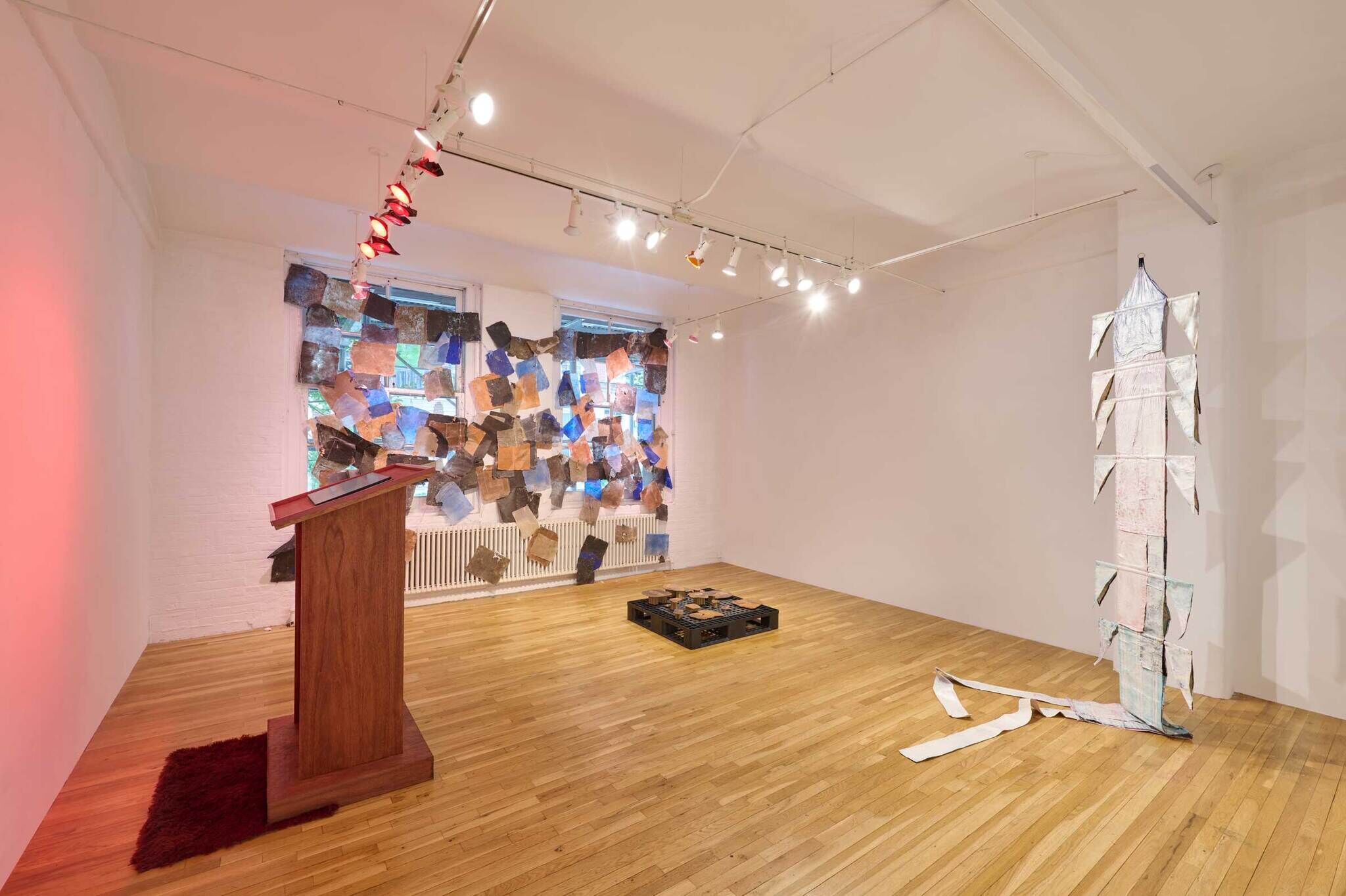 Art gallery with wooden podium, abstract wall art, floor installation, and hanging sculpture. Bright lighting and wooden flooring.