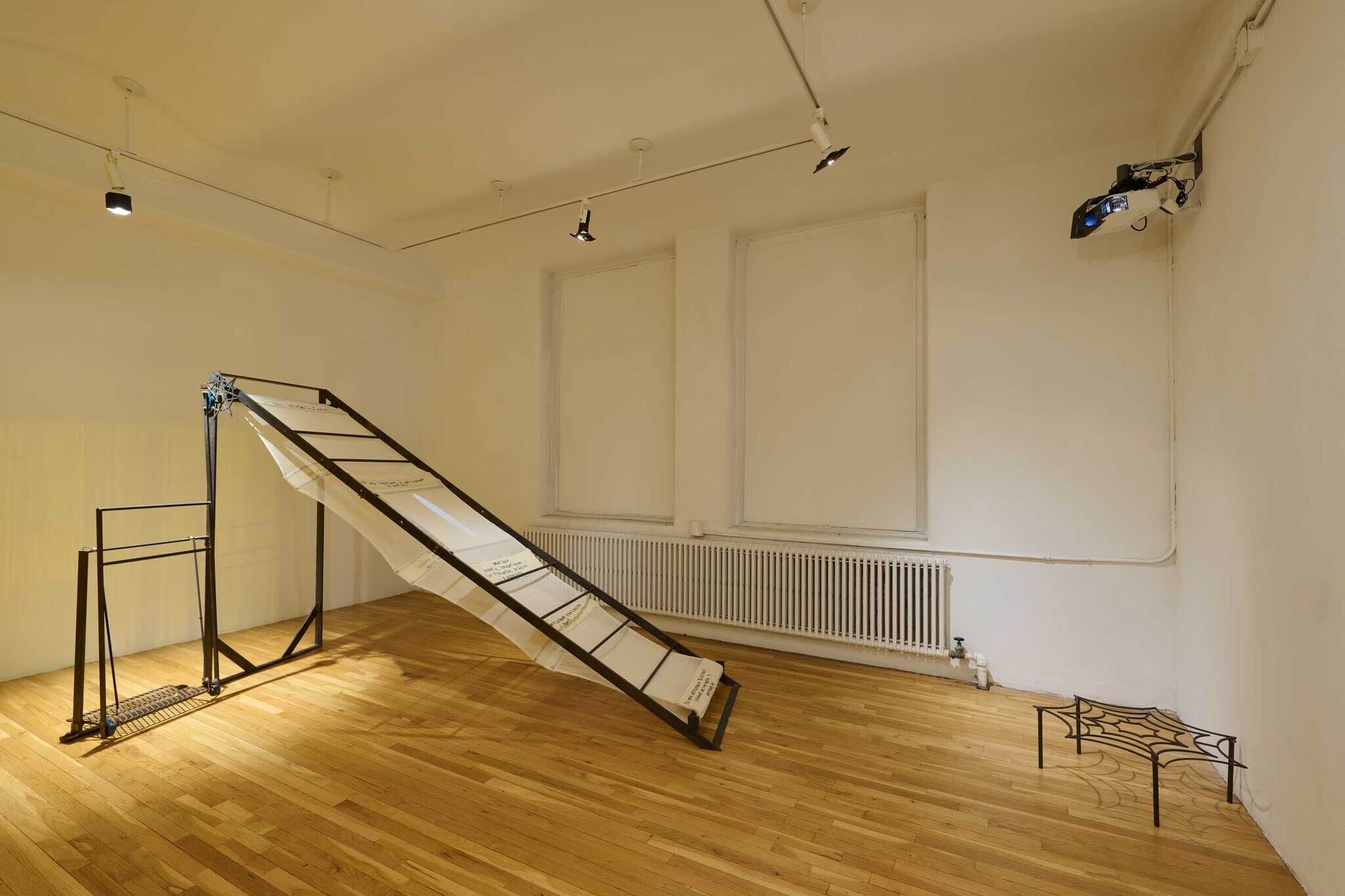 A minimalist room with wooden floors, a slanted metal structure with fabric, a small metal table, and a projector mounted on the wall.