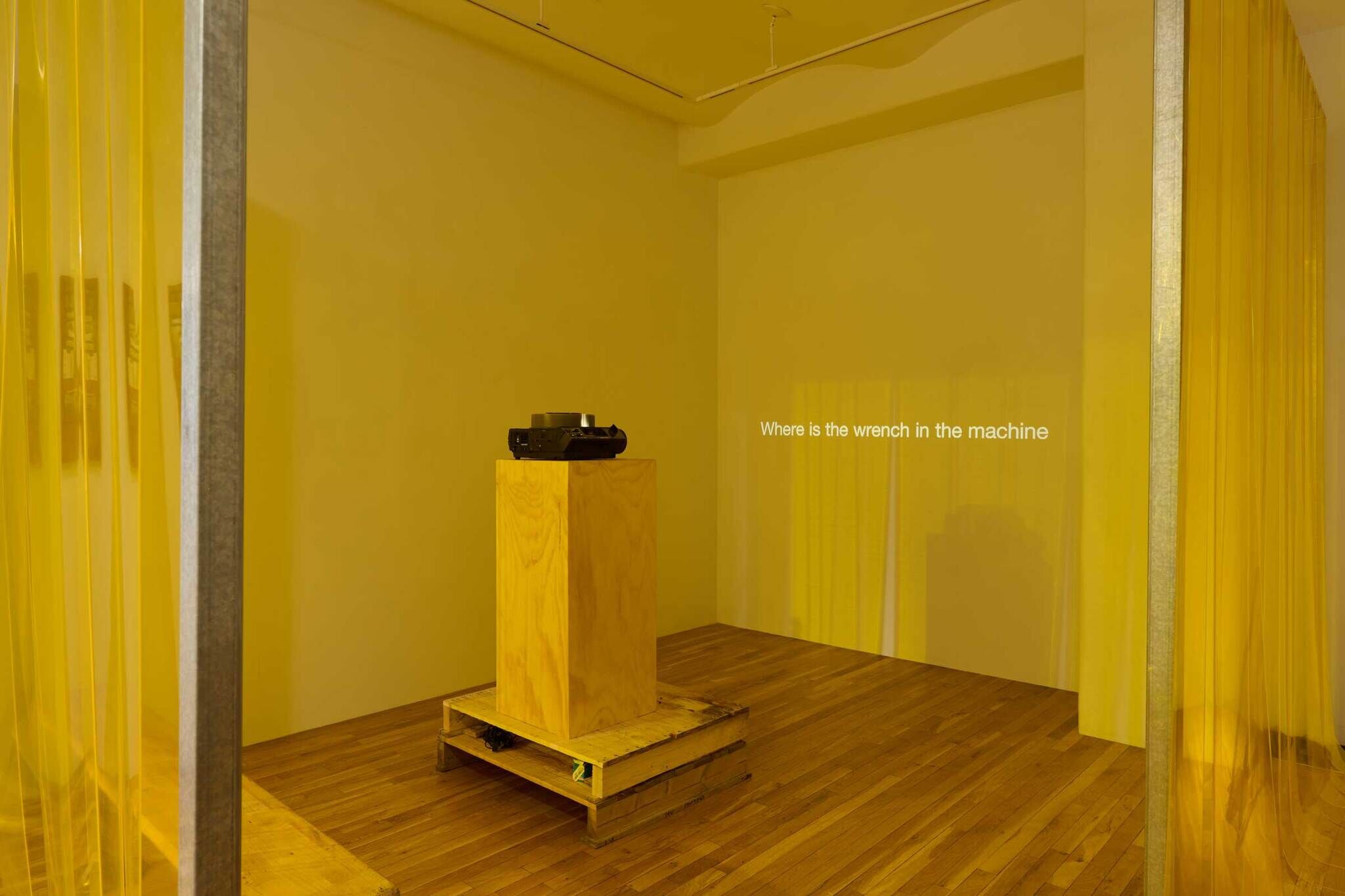 A room with yellow translucent curtains, a wooden pedestal with a projector, and the text "Where is the wrench in the machine" on the wall.