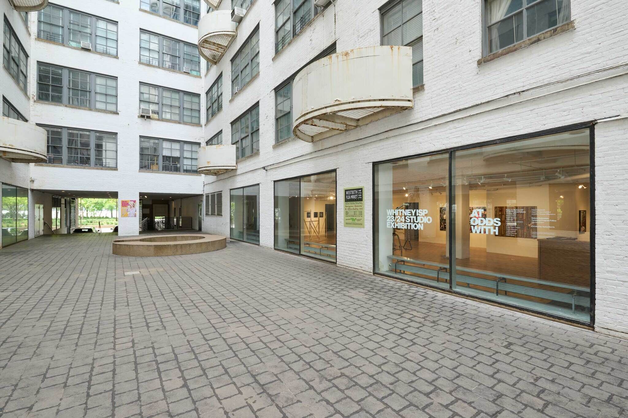 Courtyard of a white brick building with large windows, featuring an art exhibition titled "Whitney ISP 23/24 Studio Exhibition" and "At Odds With."