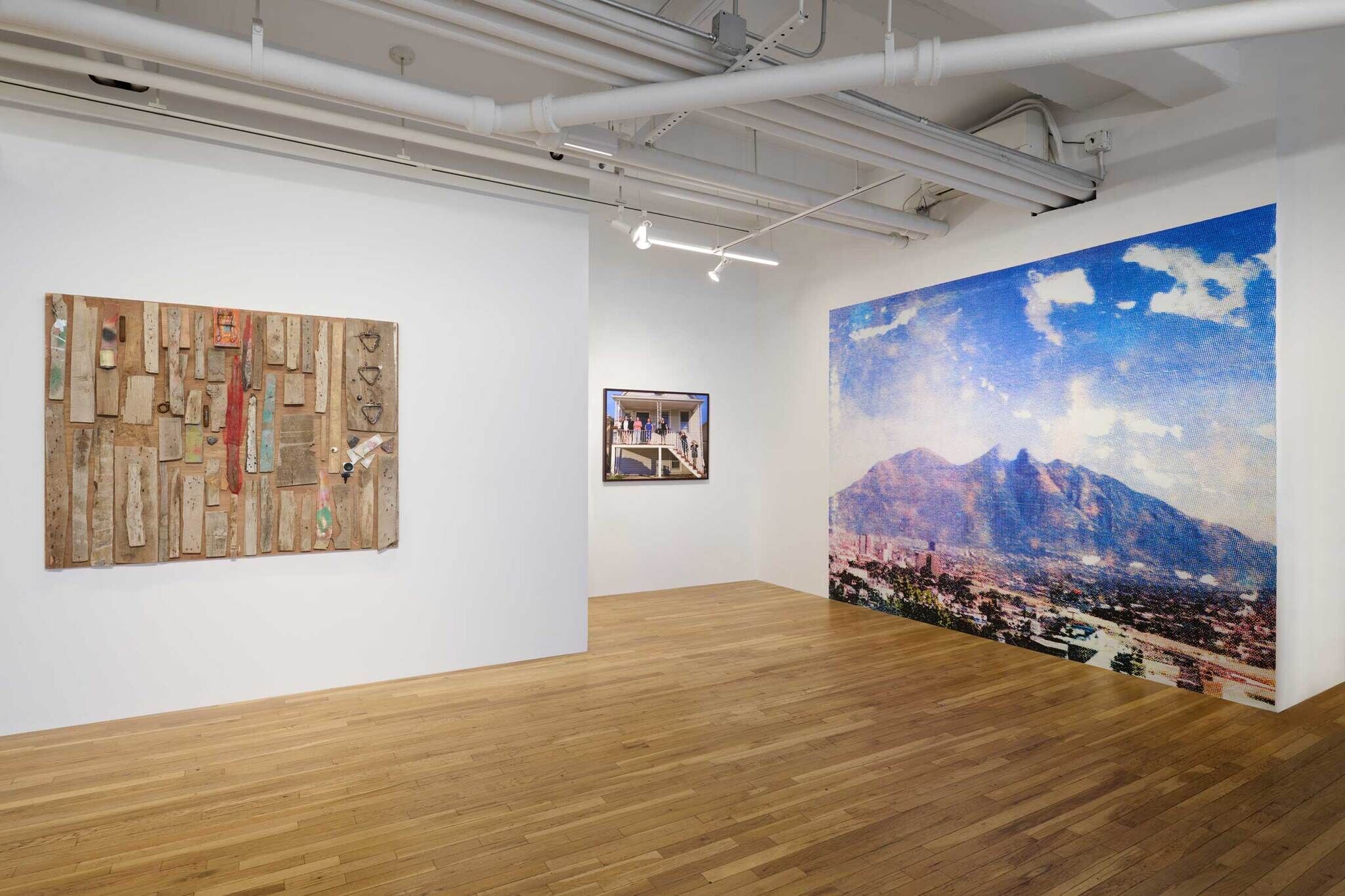 Art gallery with three pieces: a wooden collage, a photograph of people on a balcony, and a large mural of a mountain landscape.