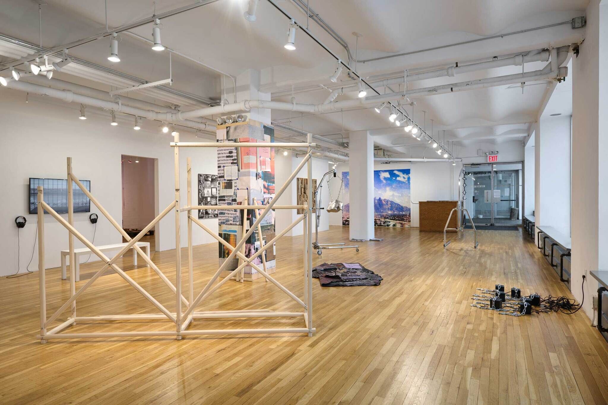 Spacious art gallery with wooden floors, various modern art installations, and bright overhead lighting. Large windows and an exit door in the background.