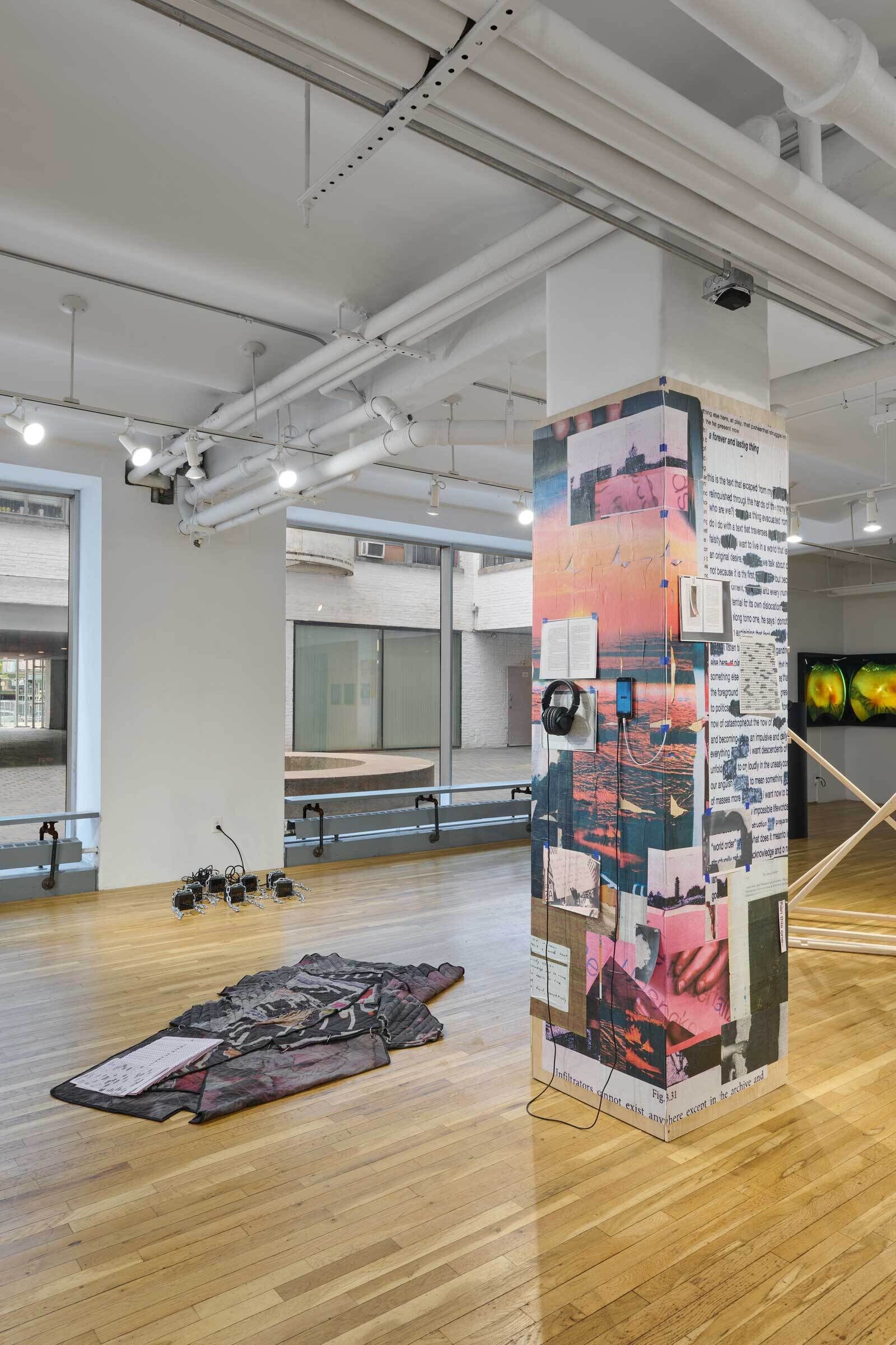 Art gallery with a pillar covered in mixed media art, a fabric piece on the wooden floor, and large windows letting in natural light.