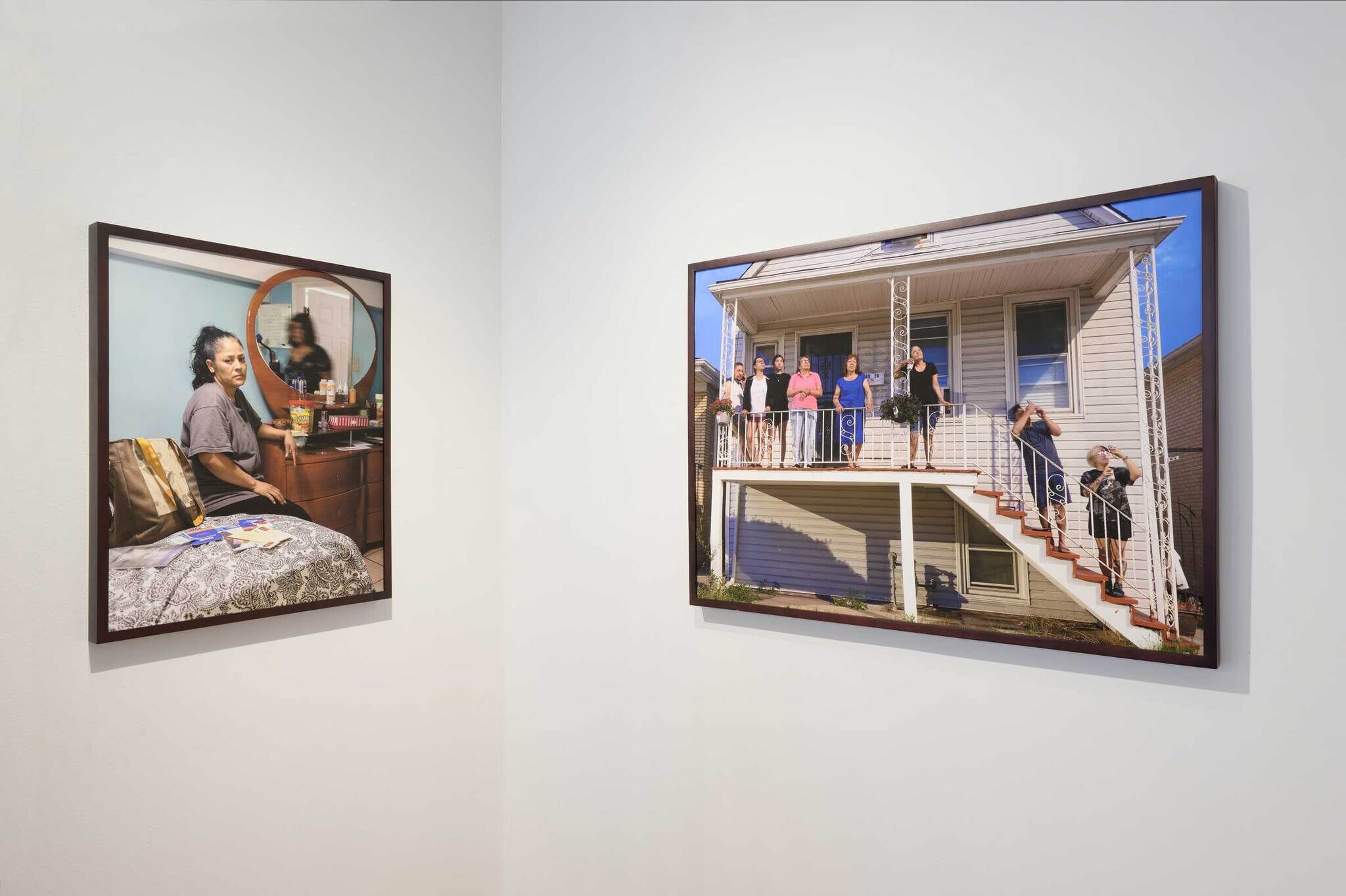 Two framed photographs on a wall: one of a woman sitting on a bed in a bedroom, and another of a group of people standing on a house balcony.