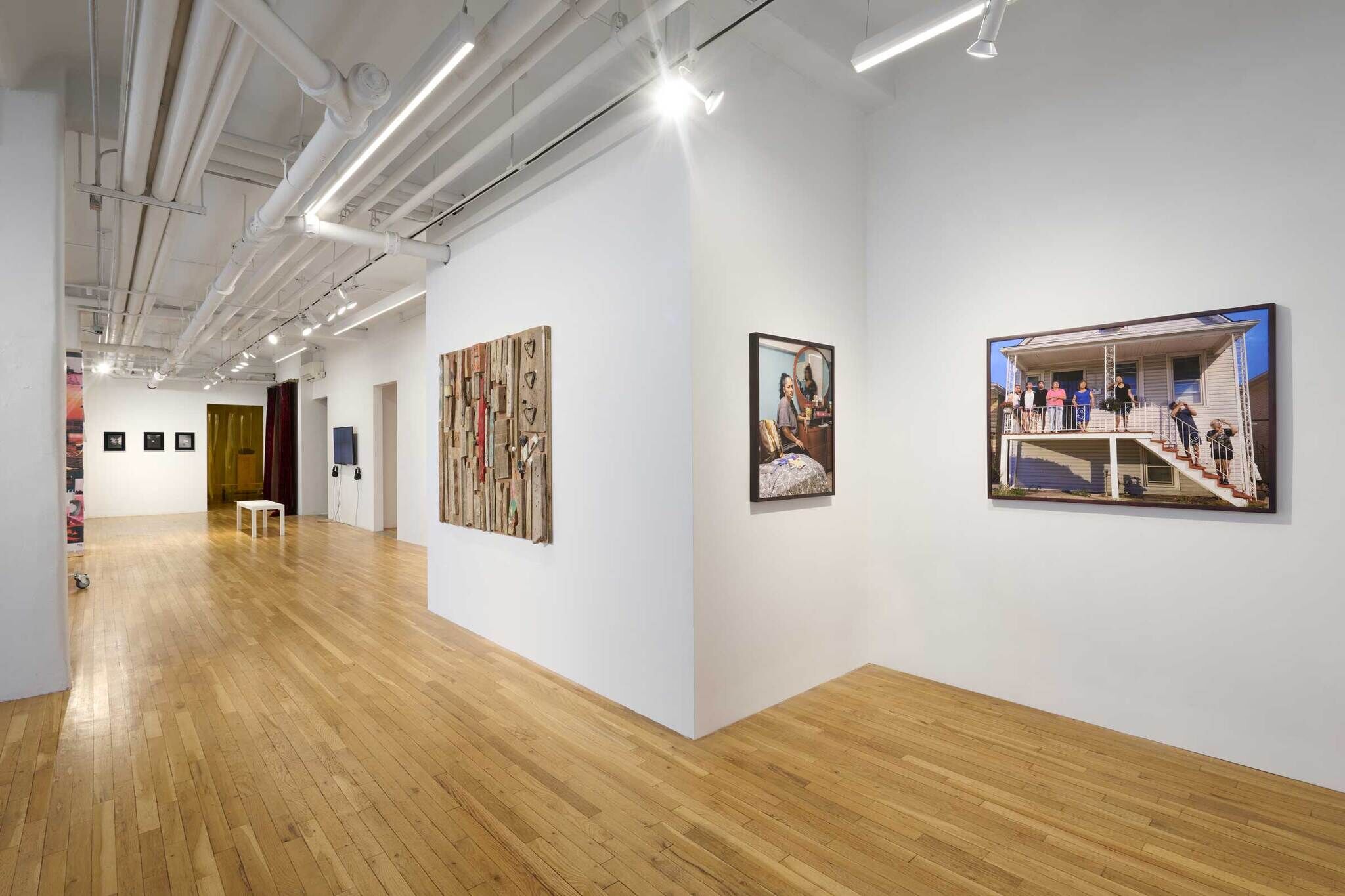 Art gallery with wooden floors, white walls, and various artworks displayed, including paintings and a mixed media piece. Bright overhead lighting.