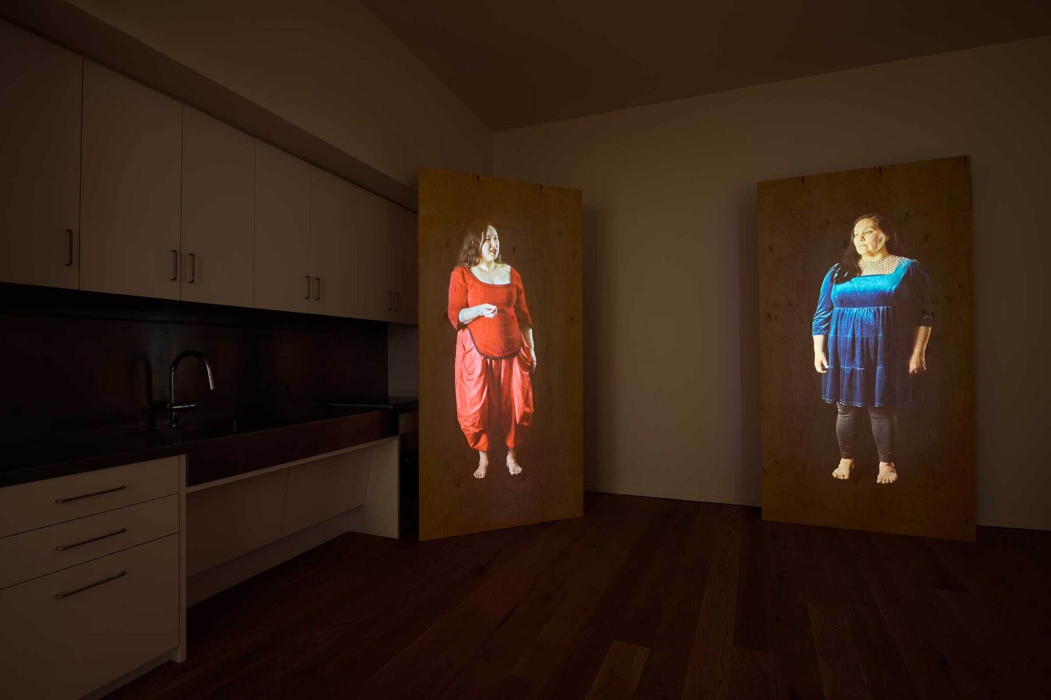 Two life-sized portraits of women, one in red and one in blue, are projected onto wooden panels in a dimly lit room with a kitchen area.