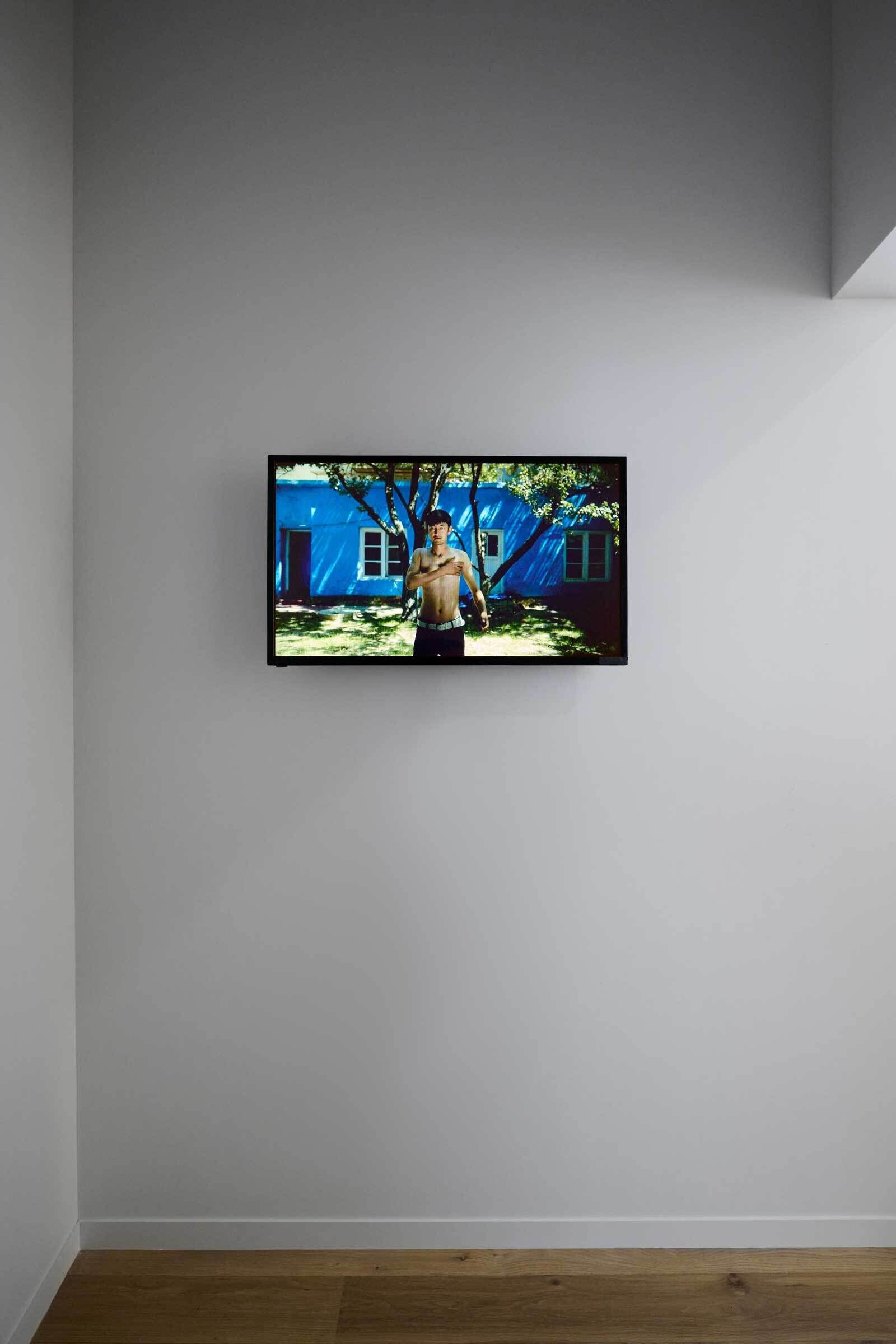 A wall-mounted TV displays a shirtless person standing in front of a blue house with trees. The room has white walls and a wooden floor.