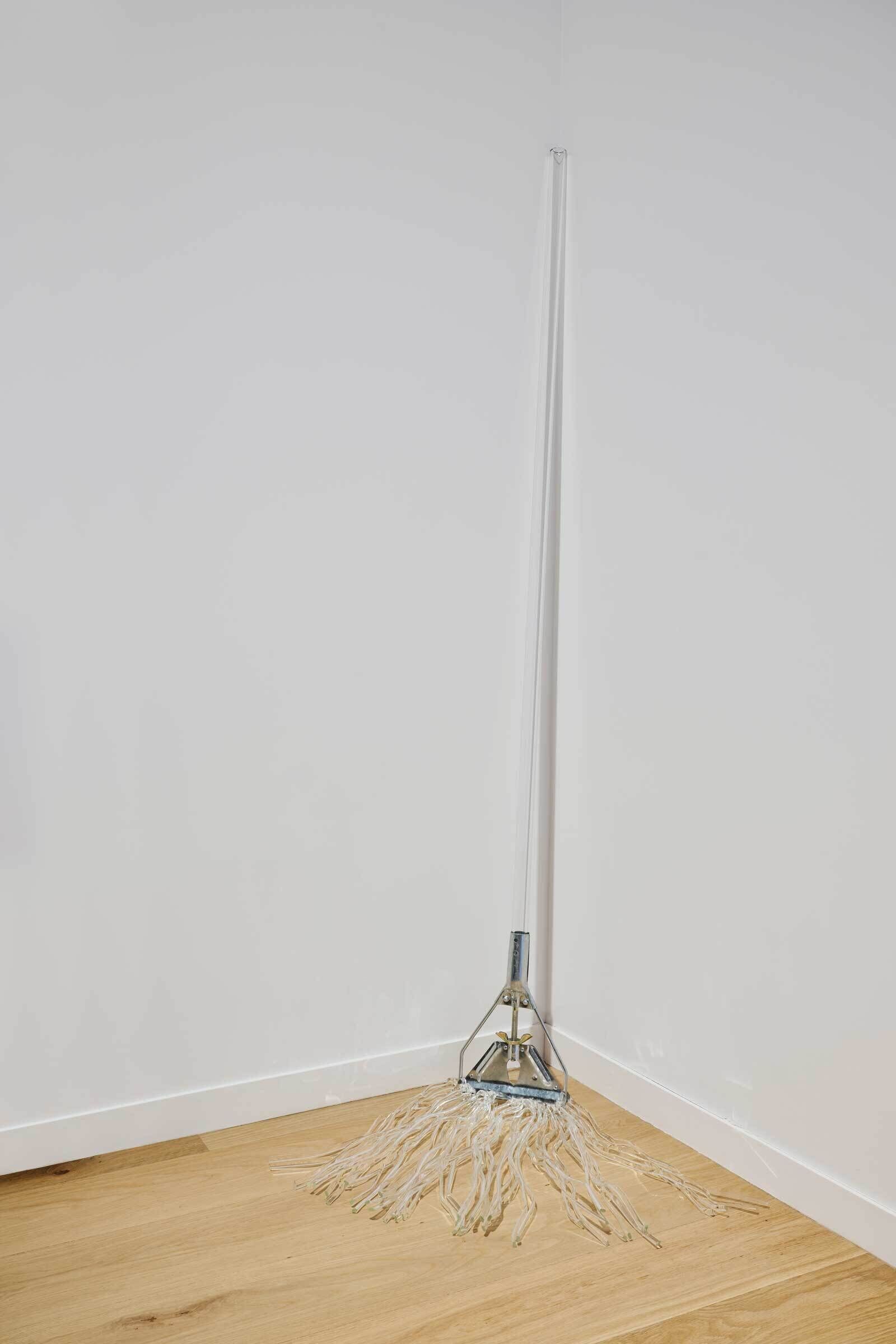 A mop with a long handle and white strands leans against the corner of a room with white walls and a wooden floor.