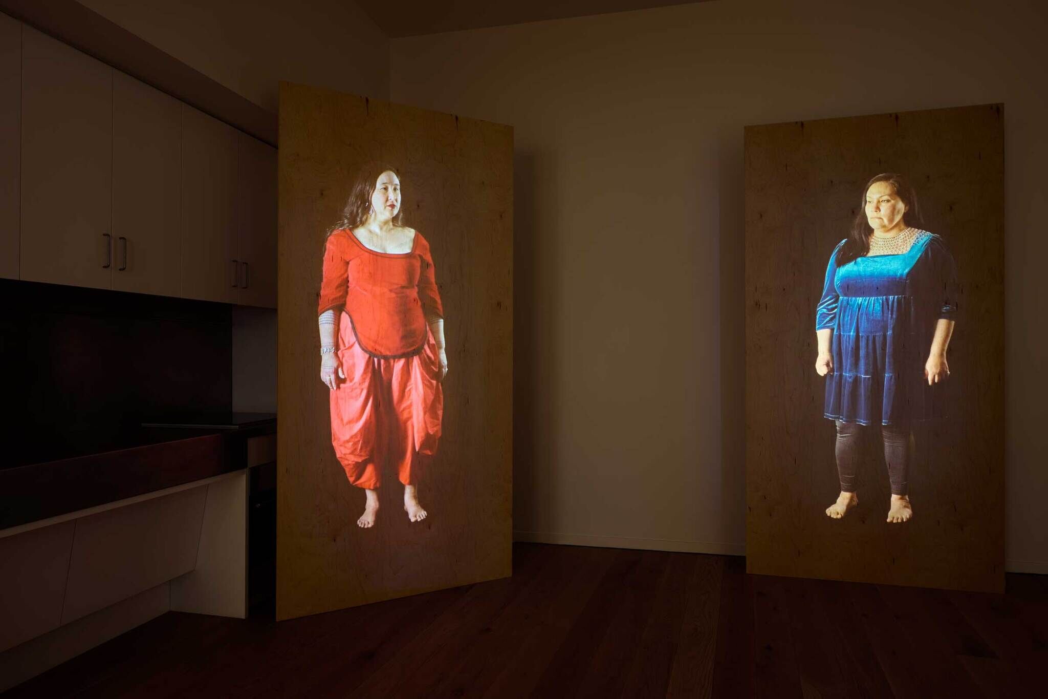 Two large wooden panels display life-sized projections of women, one in a red outfit and the other in a blue dress, in a dimly lit room.