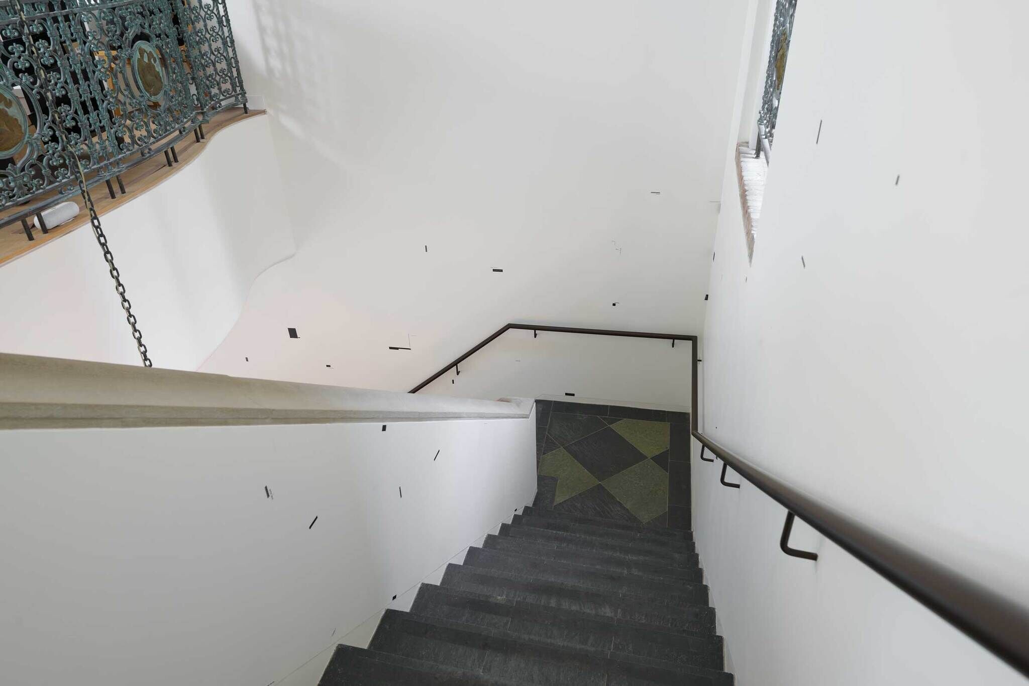 A staircase with dark steps and white walls, featuring a decorative metal railing on the left and a geometric patterned floor at the bottom.