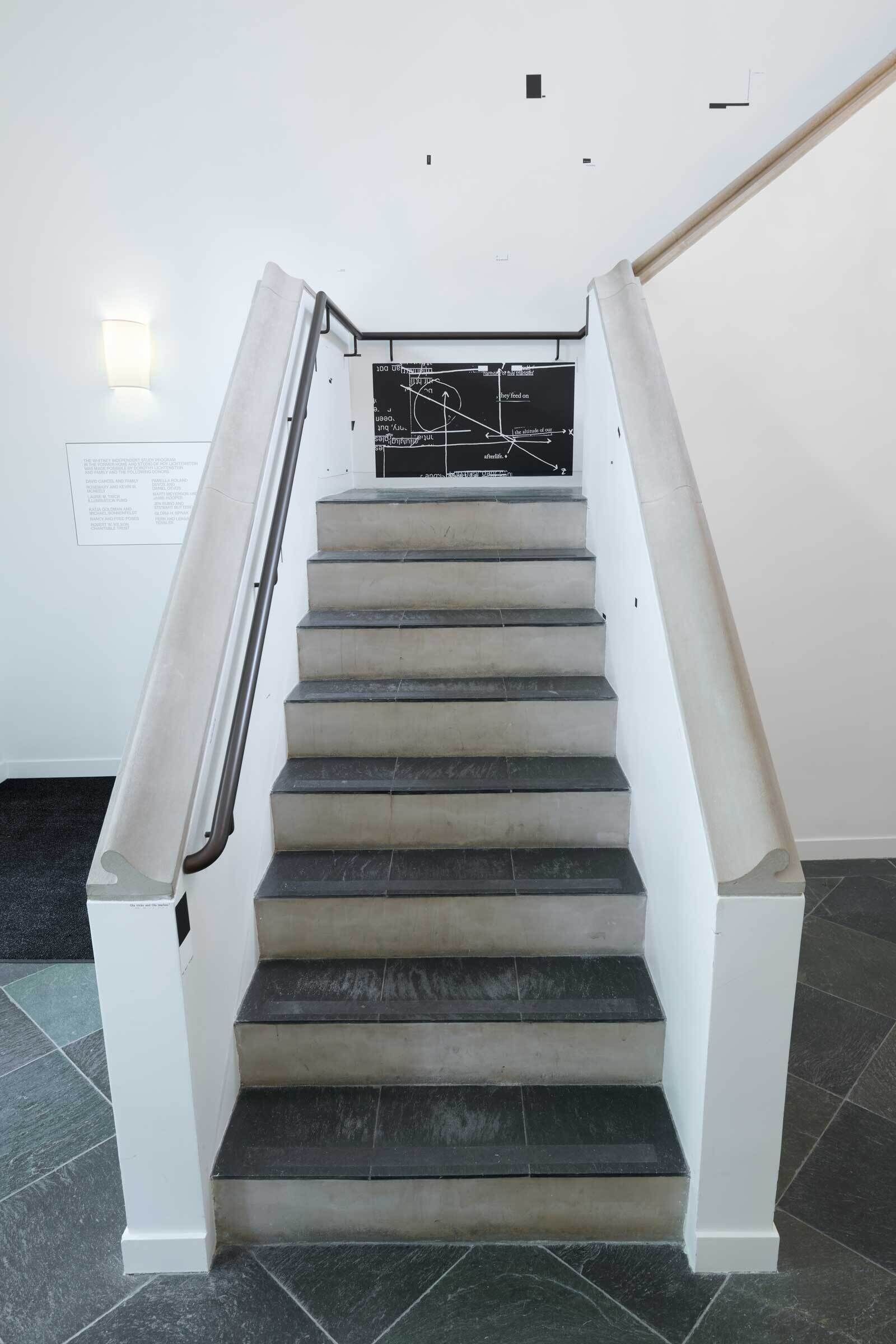 A staircase with gray steps and white railings, leading to a wall with a blackboard featuring white chalk drawings and text.