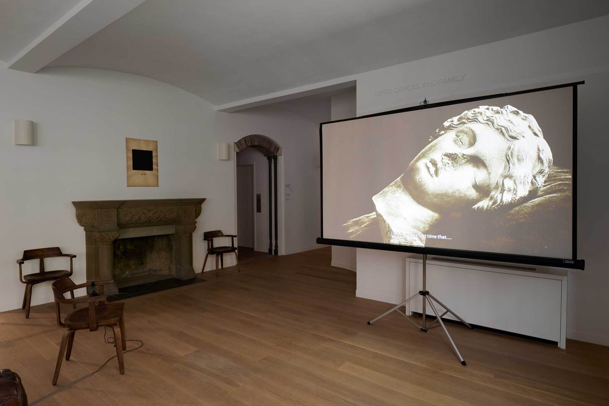 A room with wooden chairs, a fireplace, and a large screen displaying a black-and-white image of a statue's face.