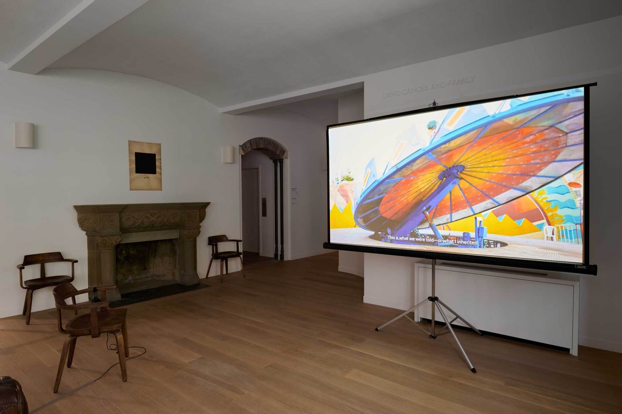 A room with a fireplace, two chairs, and a large screen displaying a colorful image of a carnival ride. Subtitles read, "This is what we were told—or what I inherited."