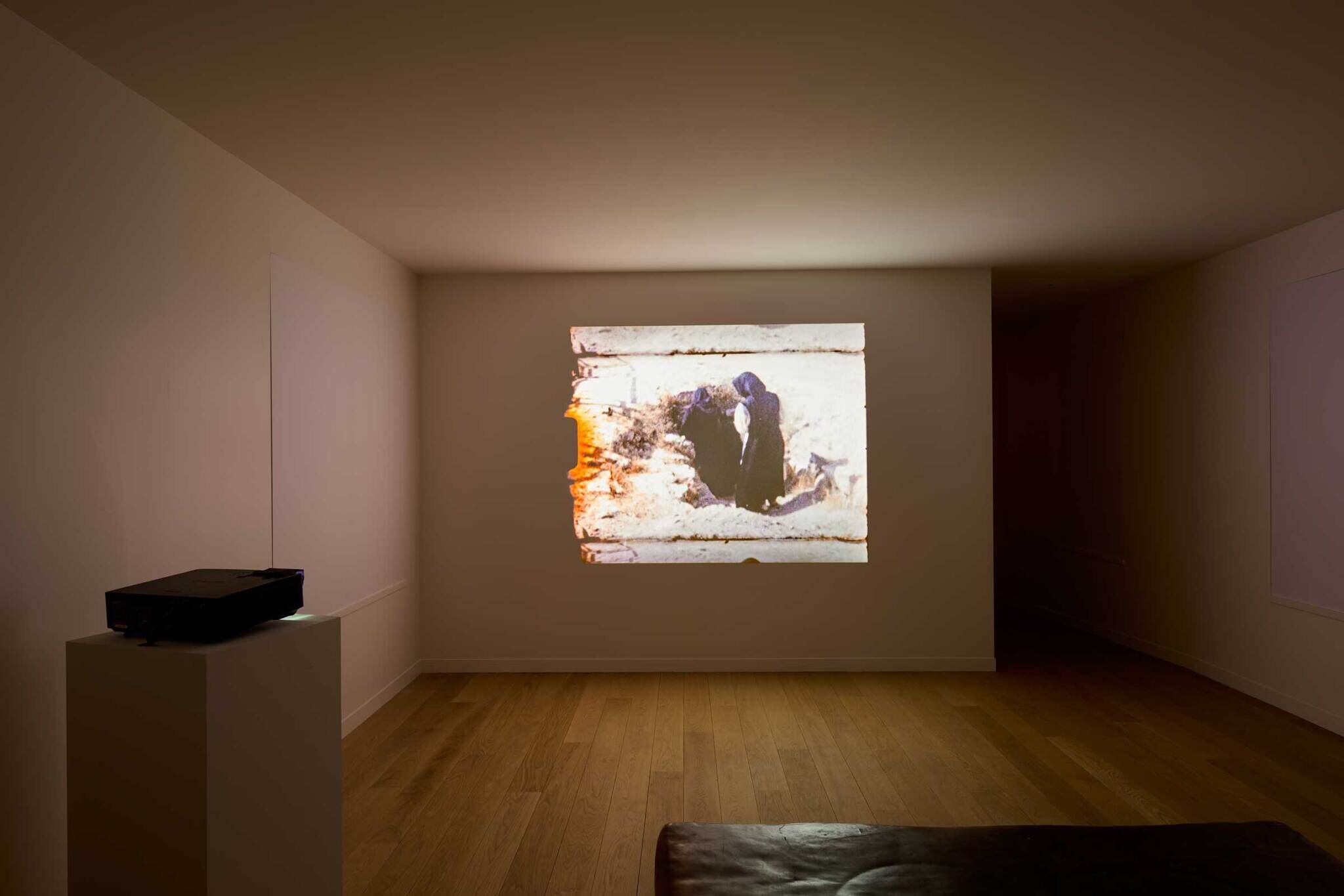 A dimly lit room with a projector displaying an old film on a white wall. The film shows two people in dark clothing outdoors.