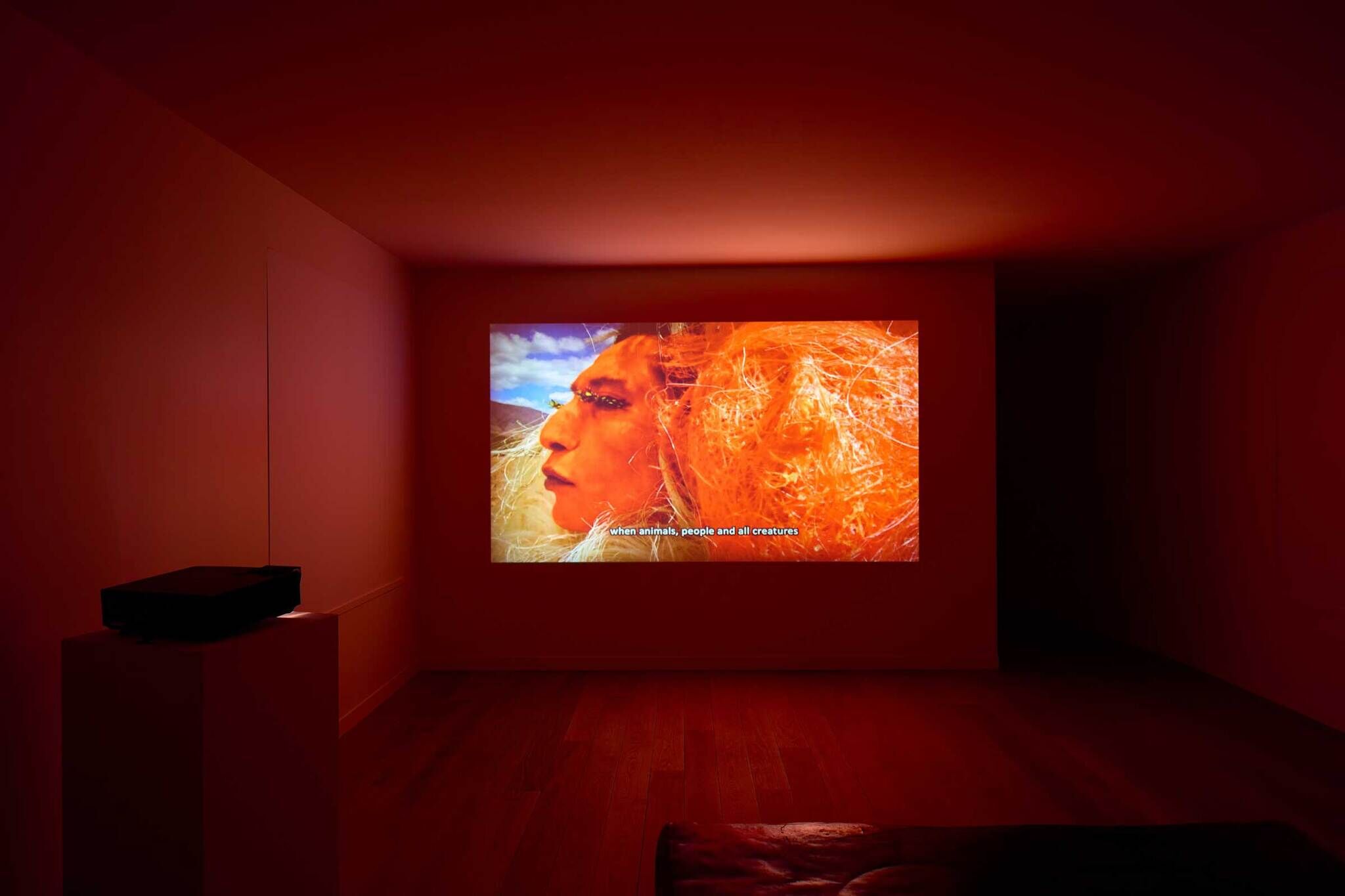 A dark room with a projector displaying a close-up of a person's face with vibrant orange and yellow hues. Subtitles read, "when animals, people and all creatures."