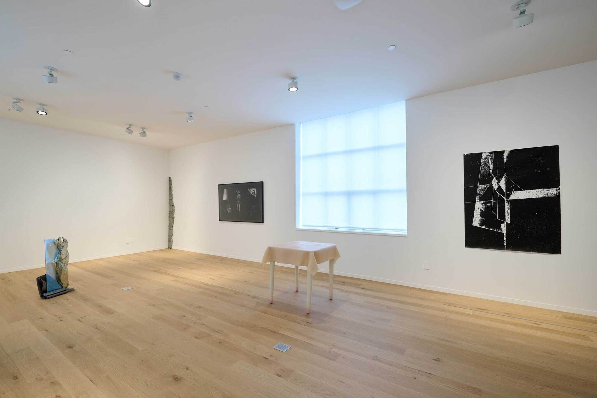 Minimalist art gallery with wooden floors, featuring abstract sculptures and paintings. A small table with a cloth is centered near a large window.