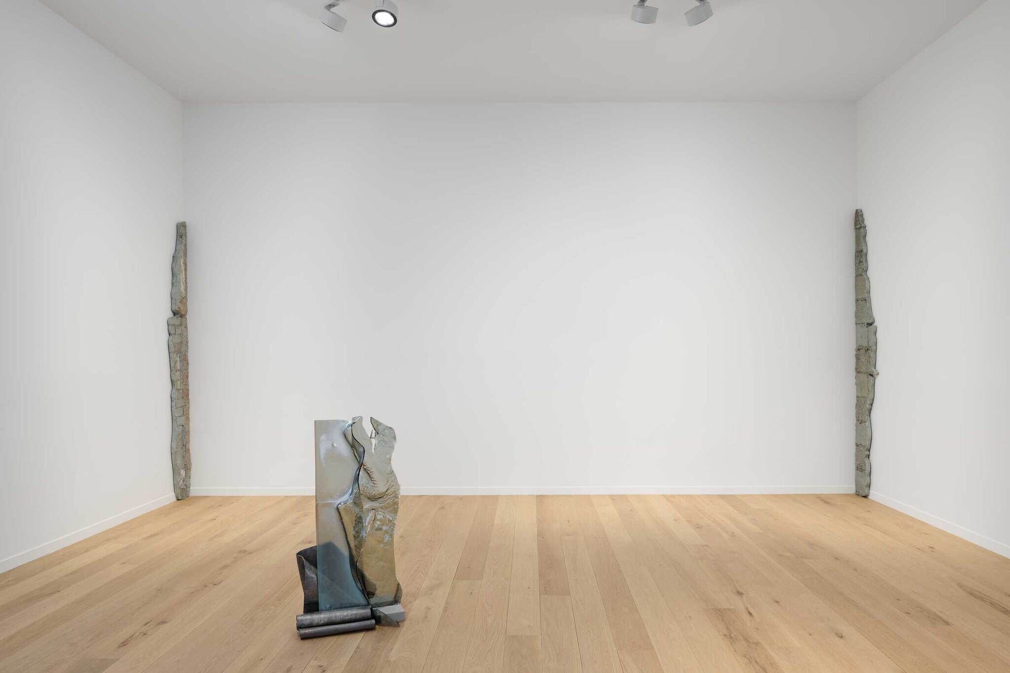 Minimalist art gallery with wooden floors, white walls, and abstract sculptures. Two tall, thin sculptures lean against the walls, and one stands on the floor.