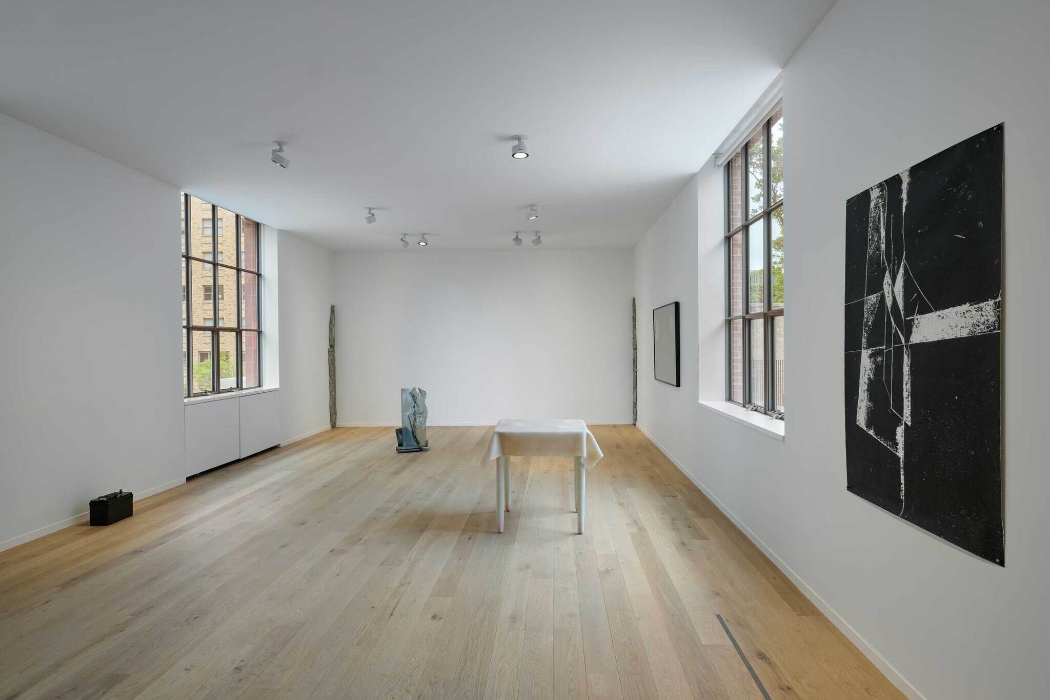 Minimalist art gallery with wooden floors, white walls, and large windows. Features abstract paintings, sculptures, and a table with a white cloth.