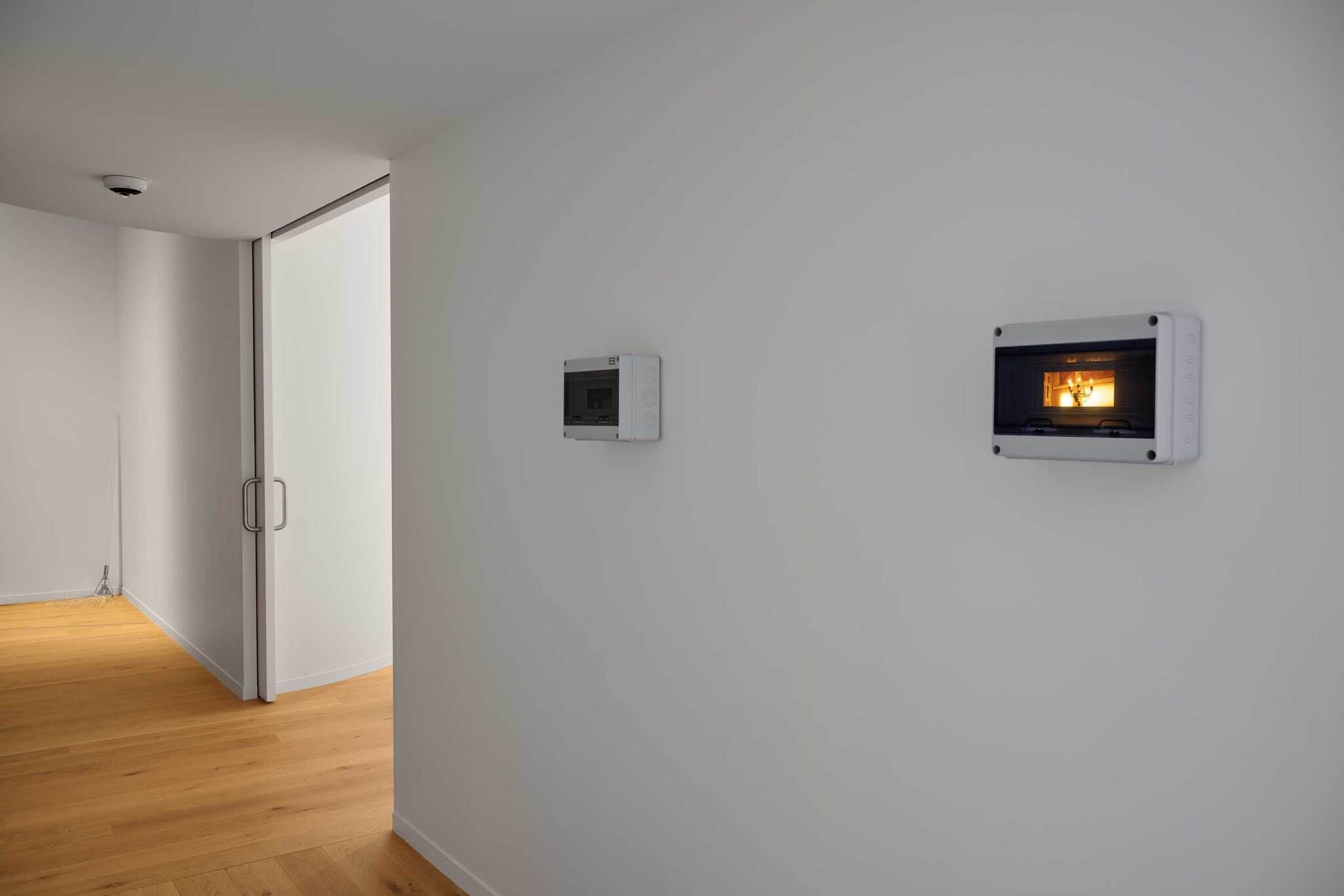 A hallway with wooden floors, white walls, a sliding door, and two electrical panels, one of which is illuminated.