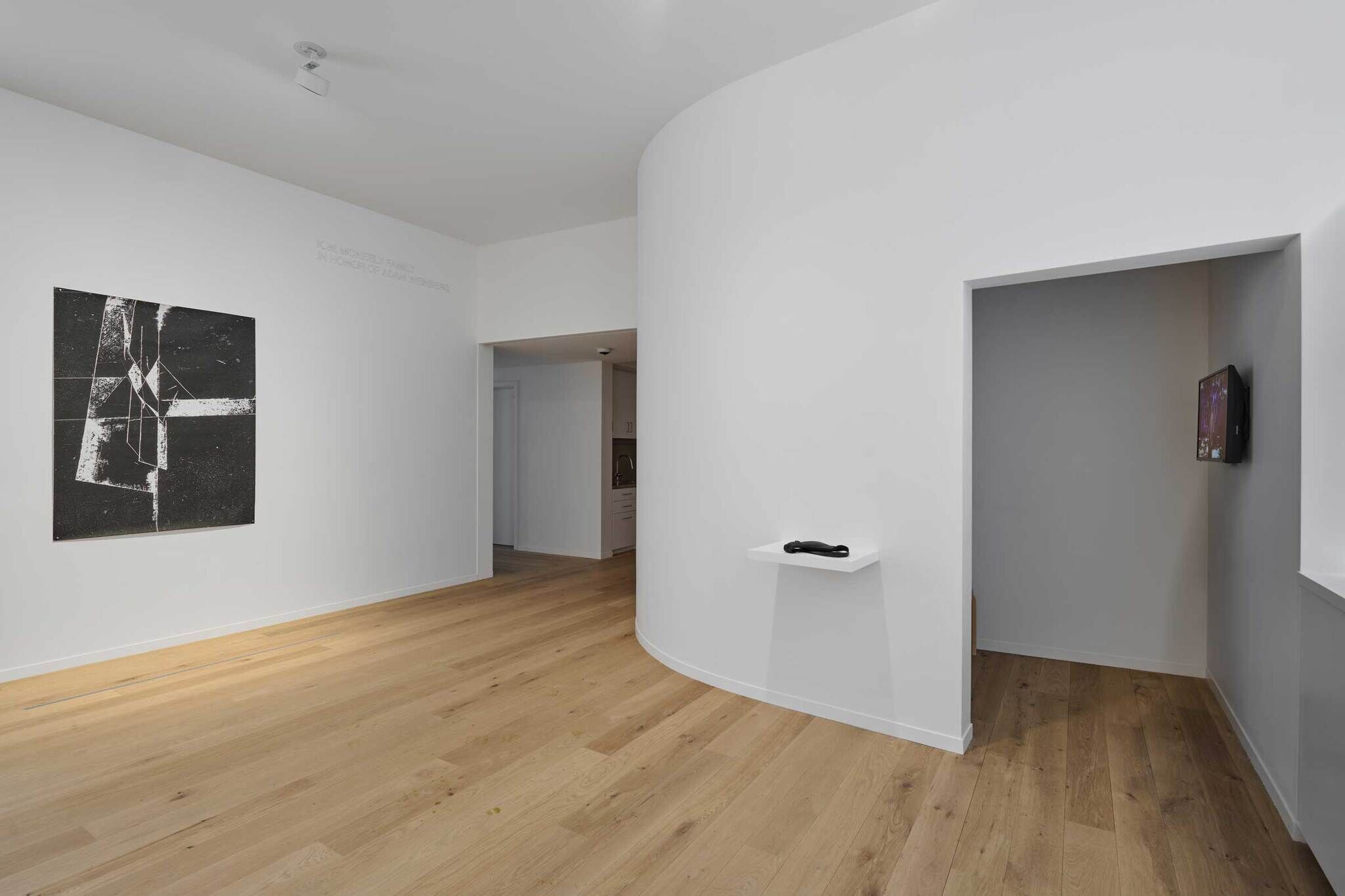 Minimalist gallery with wooden floors, white walls, abstract painting, and a small shelf holding a black object. An open doorway leads to another room.