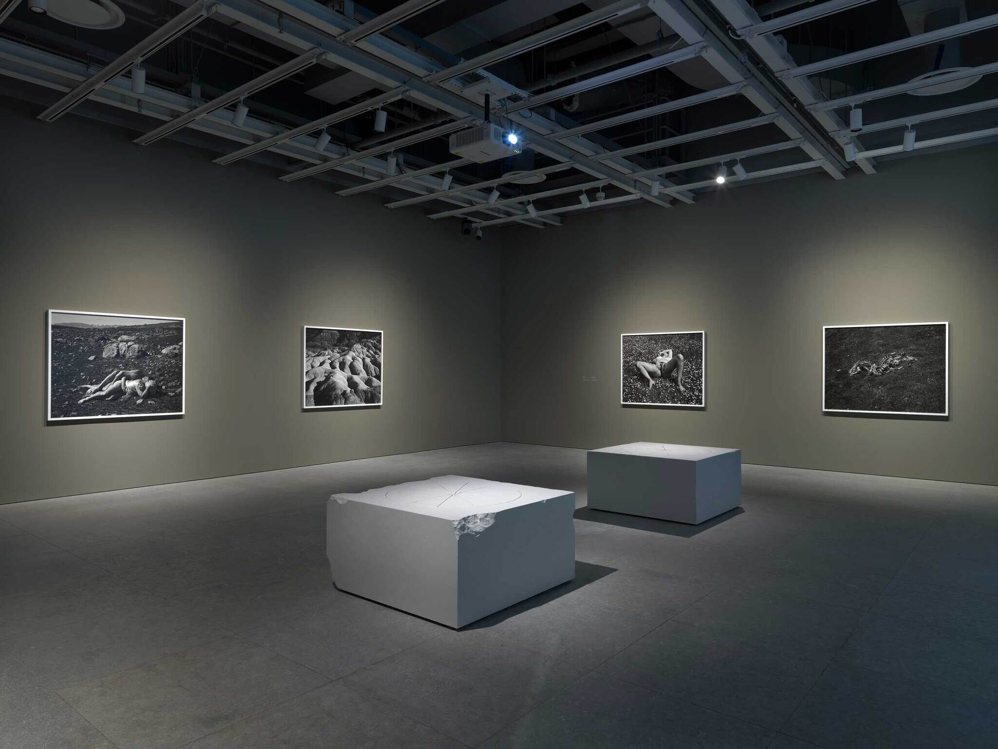 A dimly lit art gallery with five black-and-white photographs on the walls and two white rectangular sculptures in the center.