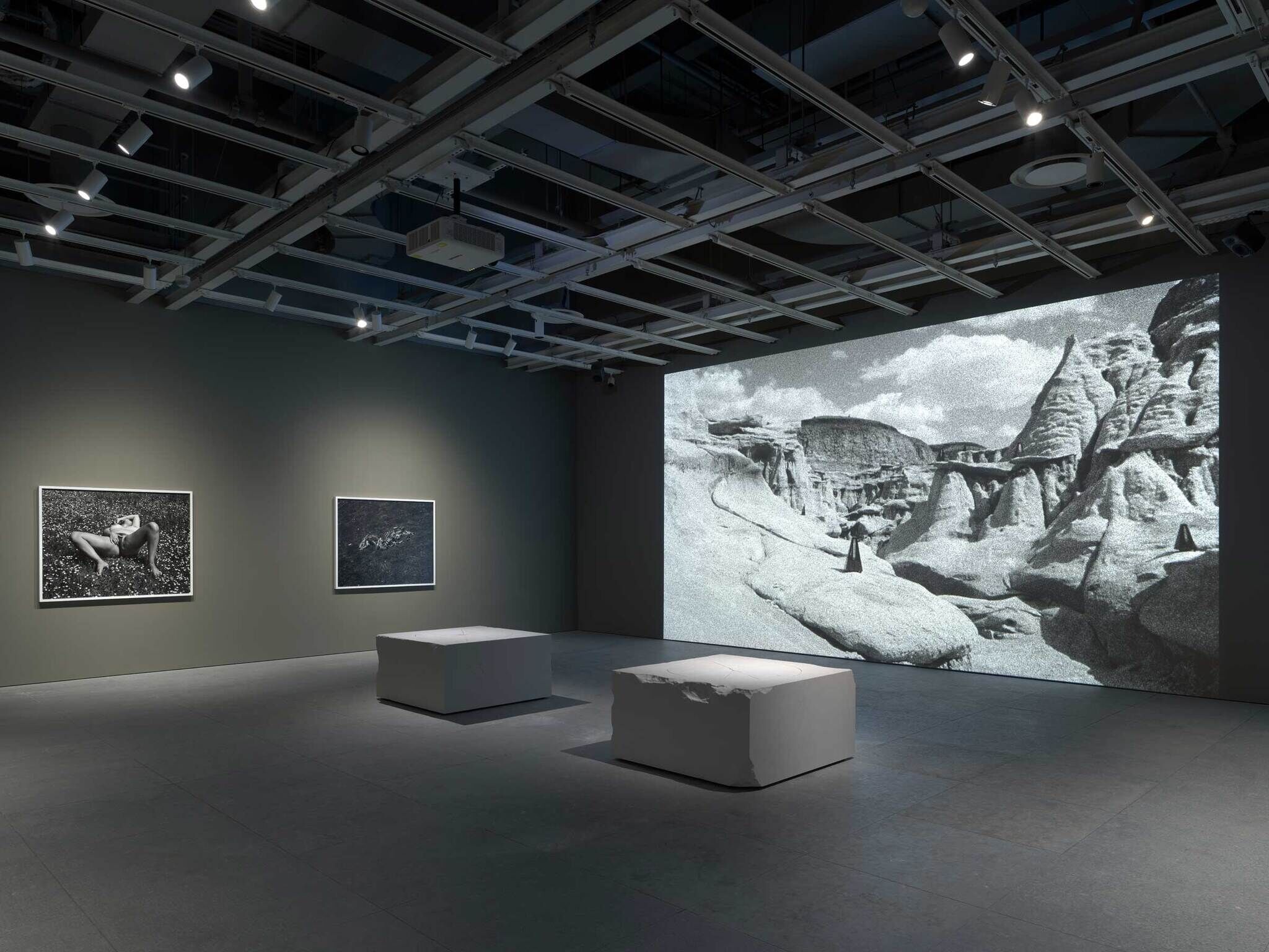 Art gallery with two framed black-and-white photos on the wall and a large projection of a rocky landscape. Two white benches in the center.