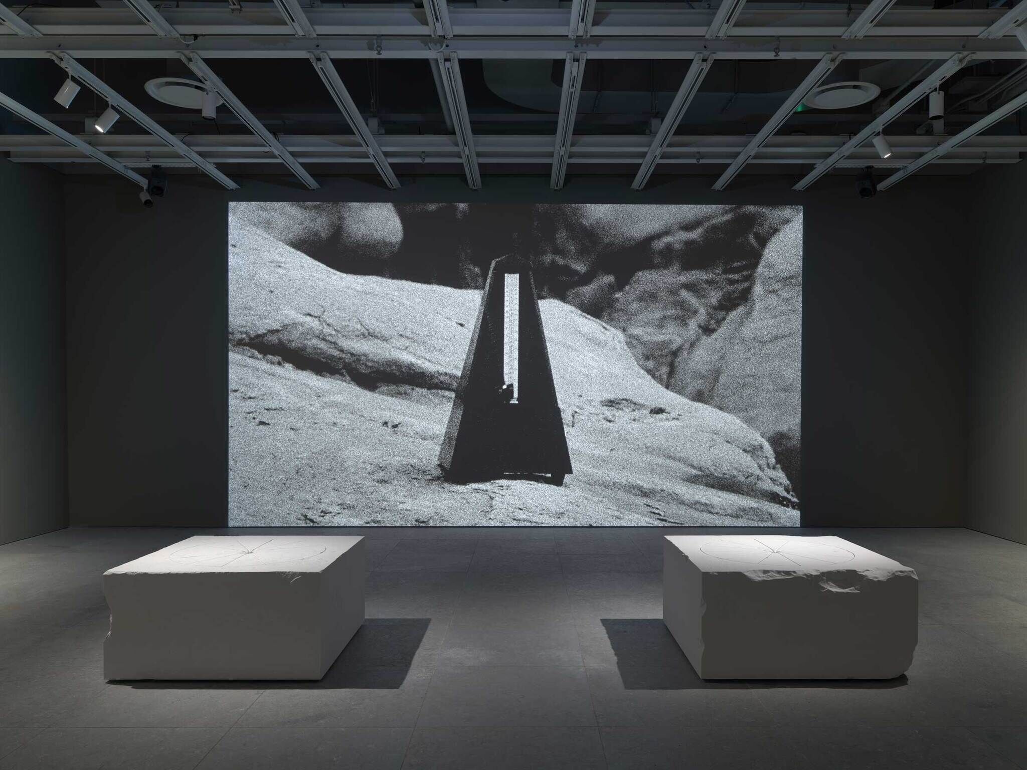 A dark room with a large black-and-white projection of a metronome on a rocky surface. Two white rectangular blocks are placed in front.