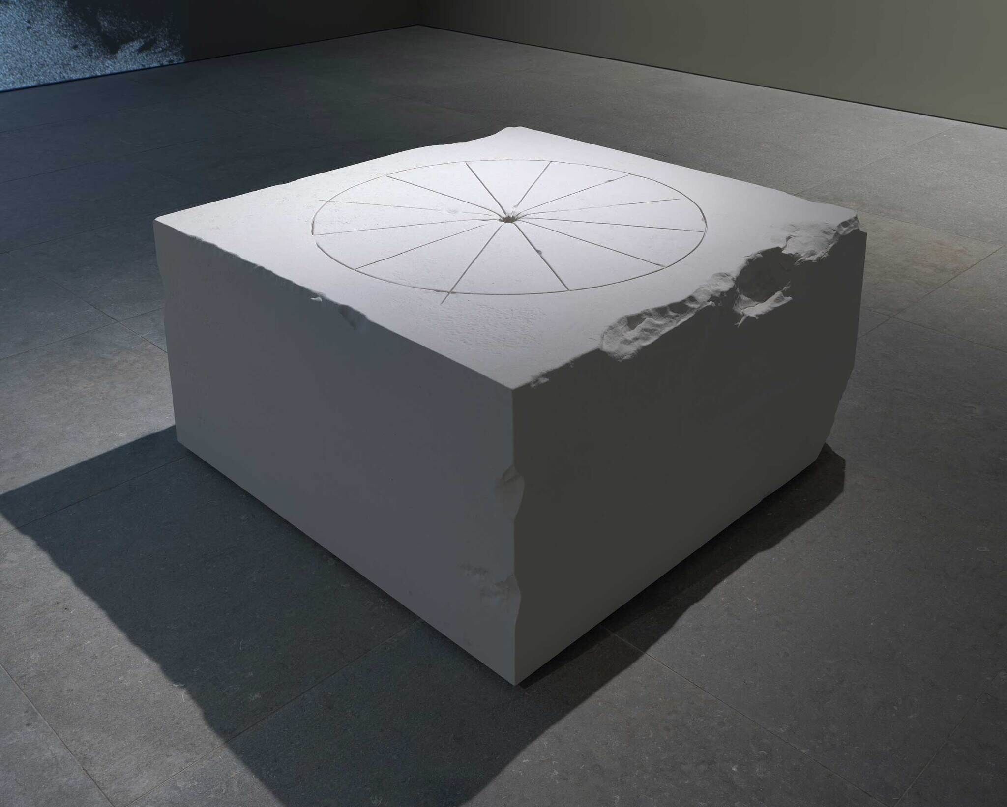 A large, white, rectangular stone block with a circular pattern etched on top, placed on a gray tiled floor in a dimly lit room.