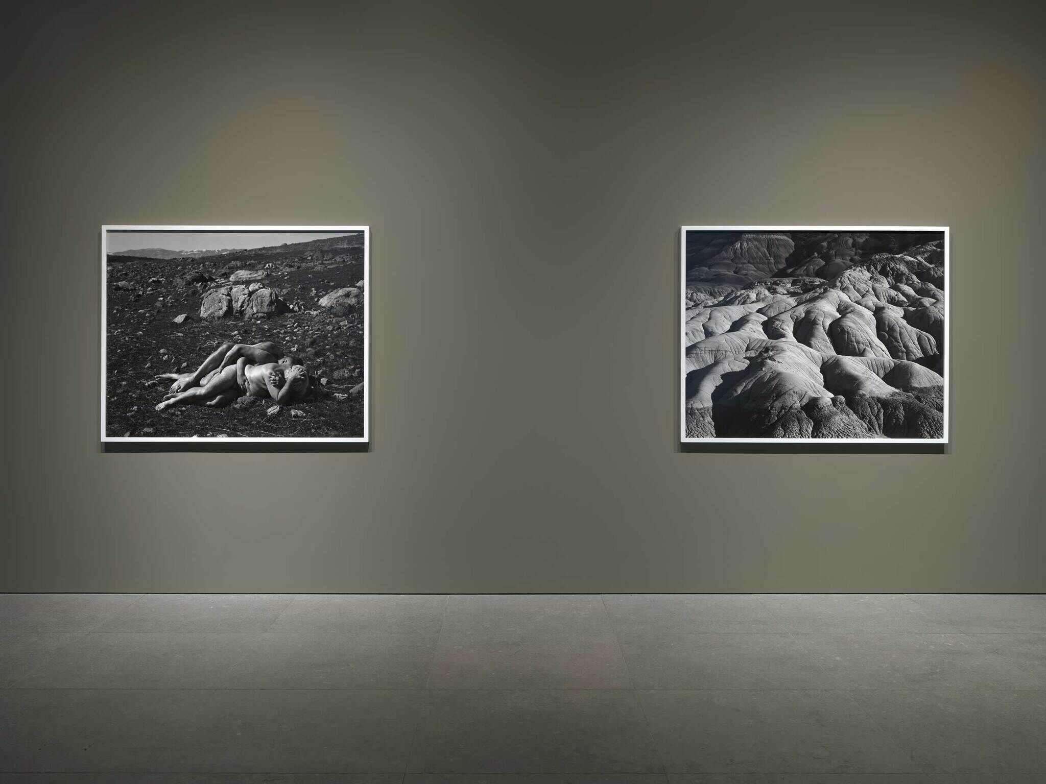 Two framed black-and-white photographs hang on a gray wall in an art gallery. The left photo shows a person lying on rocky ground, the right depicts a rugged landscape.