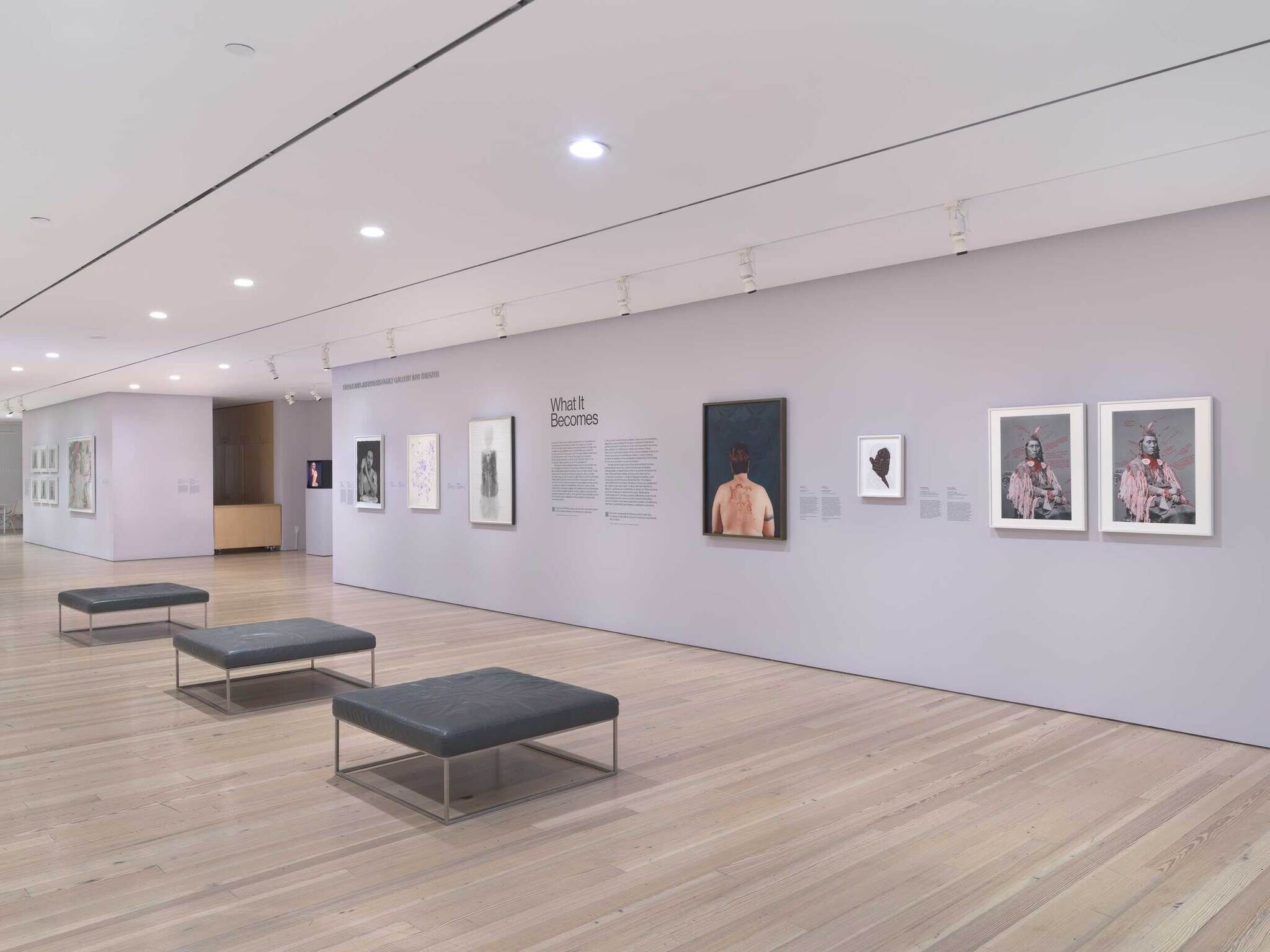 Modern art gallery with light wood floors, gray walls, and various artworks displayed. Three black benches are placed in the center.