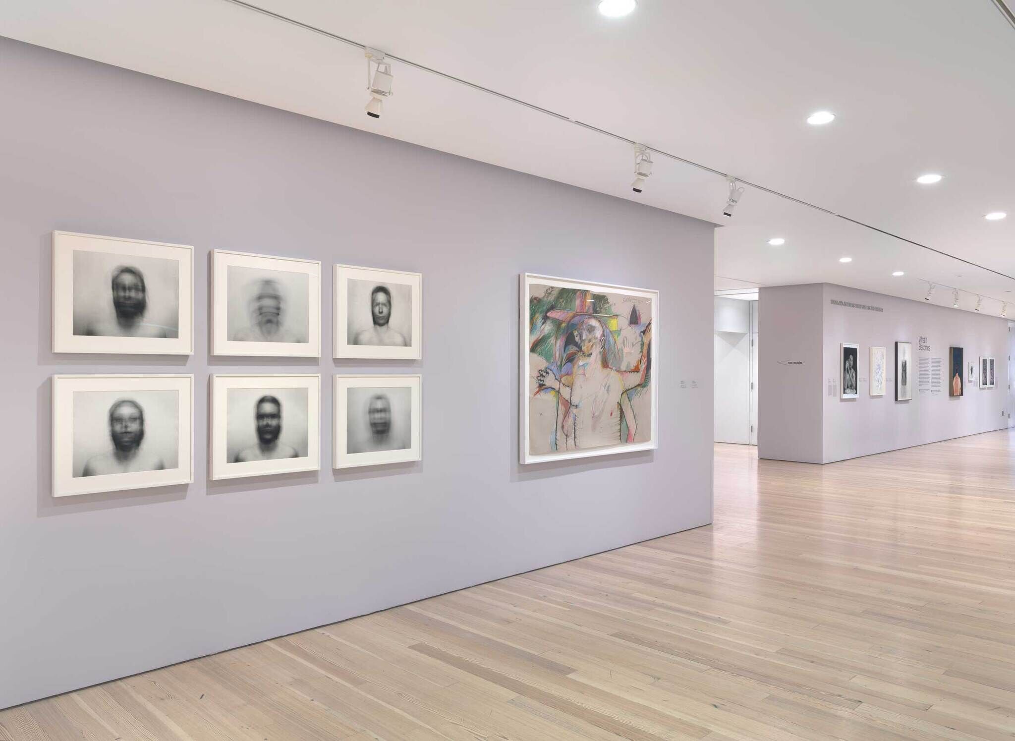 Art gallery with six blurred portraits and a colorful abstract painting on a light gray wall, wooden floor, and additional artworks in the background.