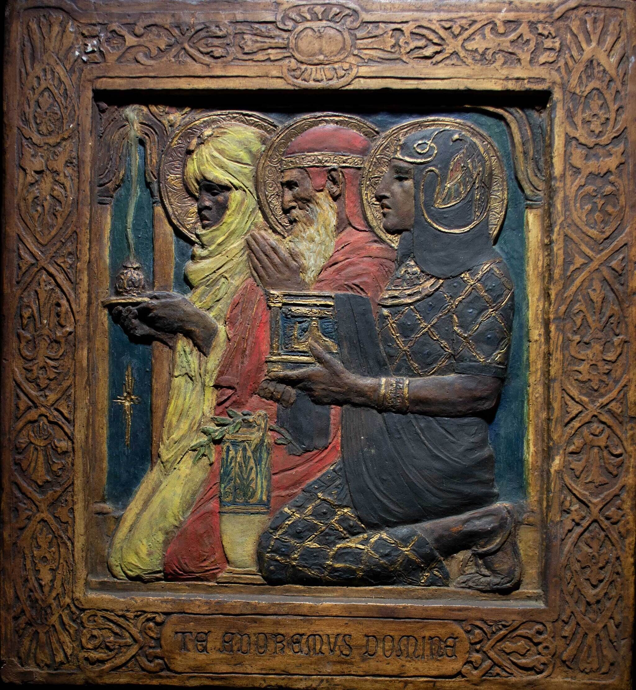 A carved and painted relief depicting three figures in ornate attire, each holding a gift, with intricate border designs and Latin text below.