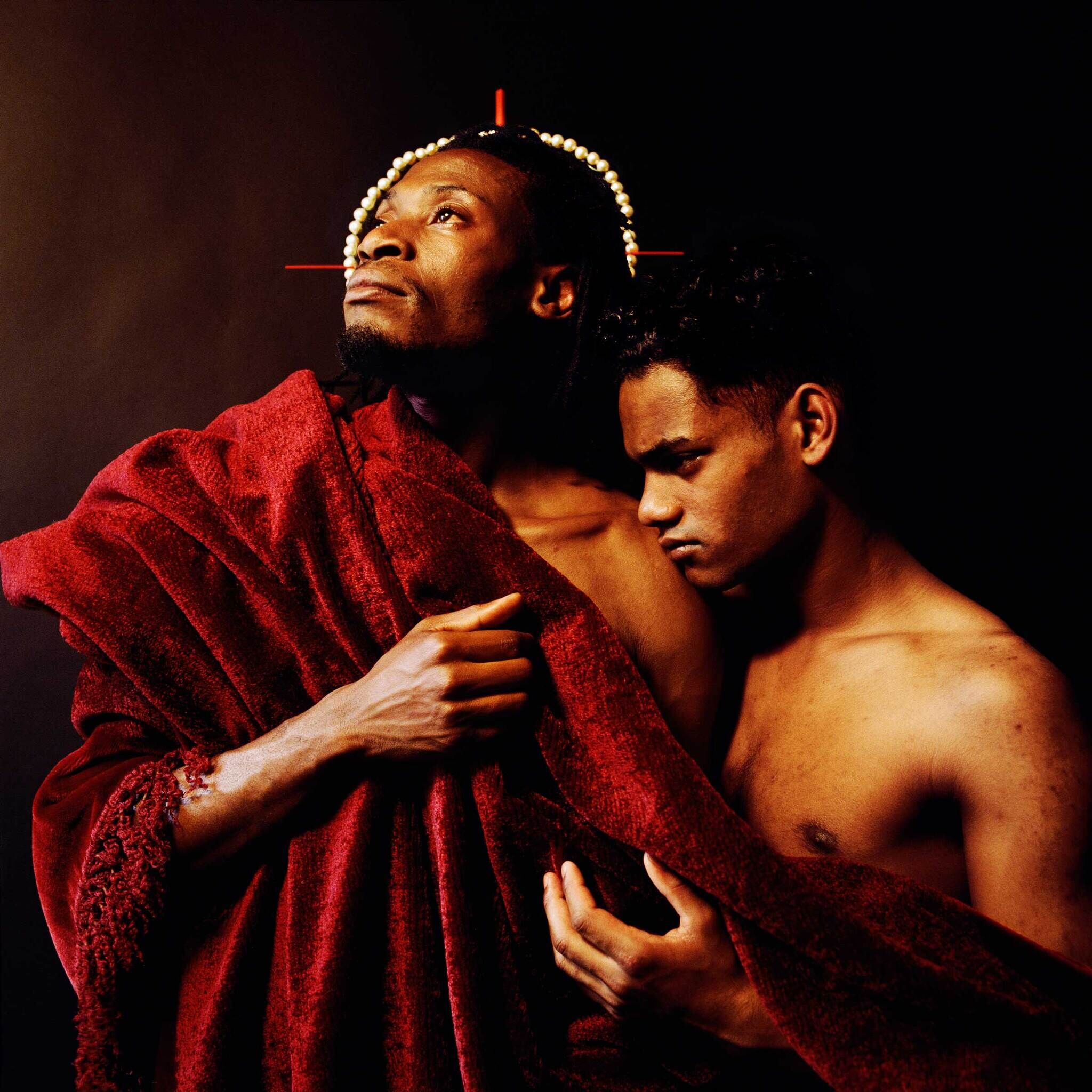 Two shirtless individuals draped in a red cloth, one looking upward with a halo-like headpiece, the other looking downward, both against a dark background.