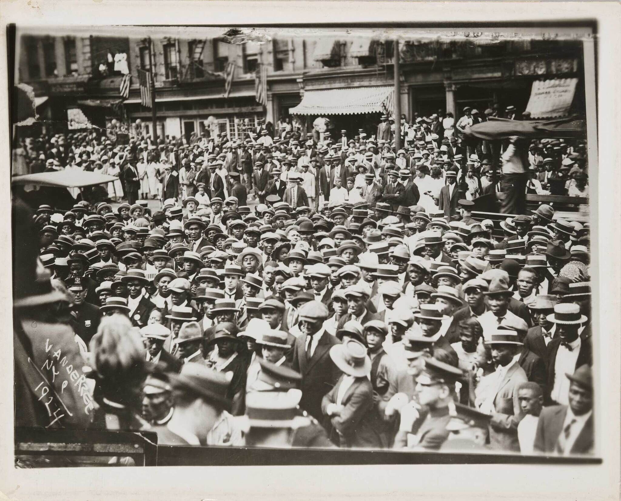 A large crowd of people, mostly wearing hats, gathered on a city street with buildings and American flags in the background.