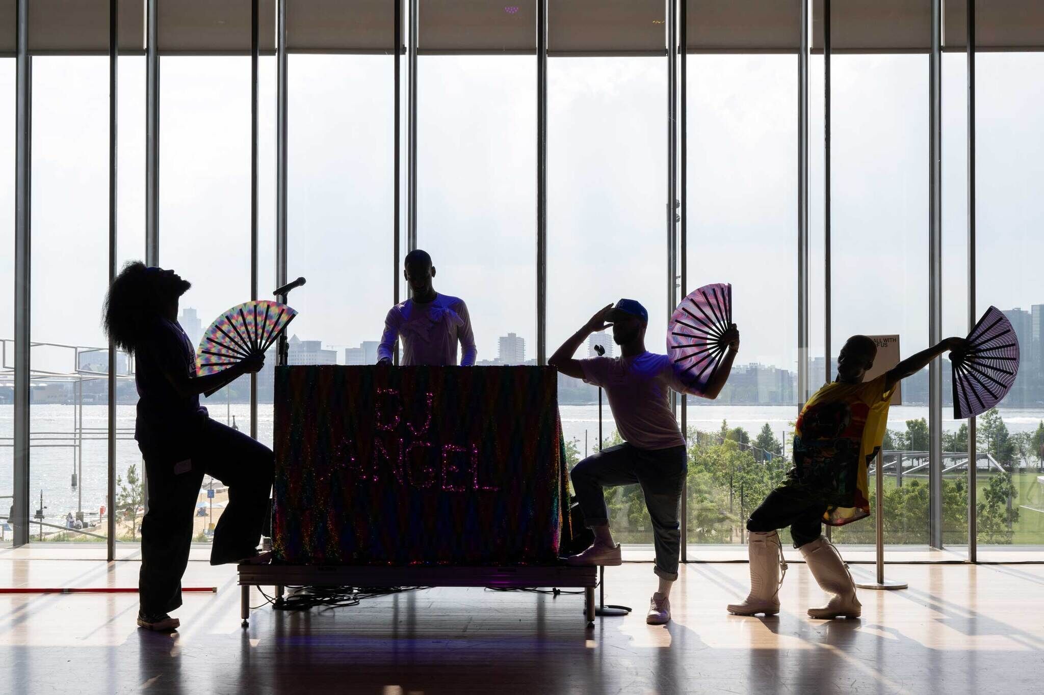 Silhouetted performers with fans dance around a DJ in front of large windows overlooking a cityscape and river.