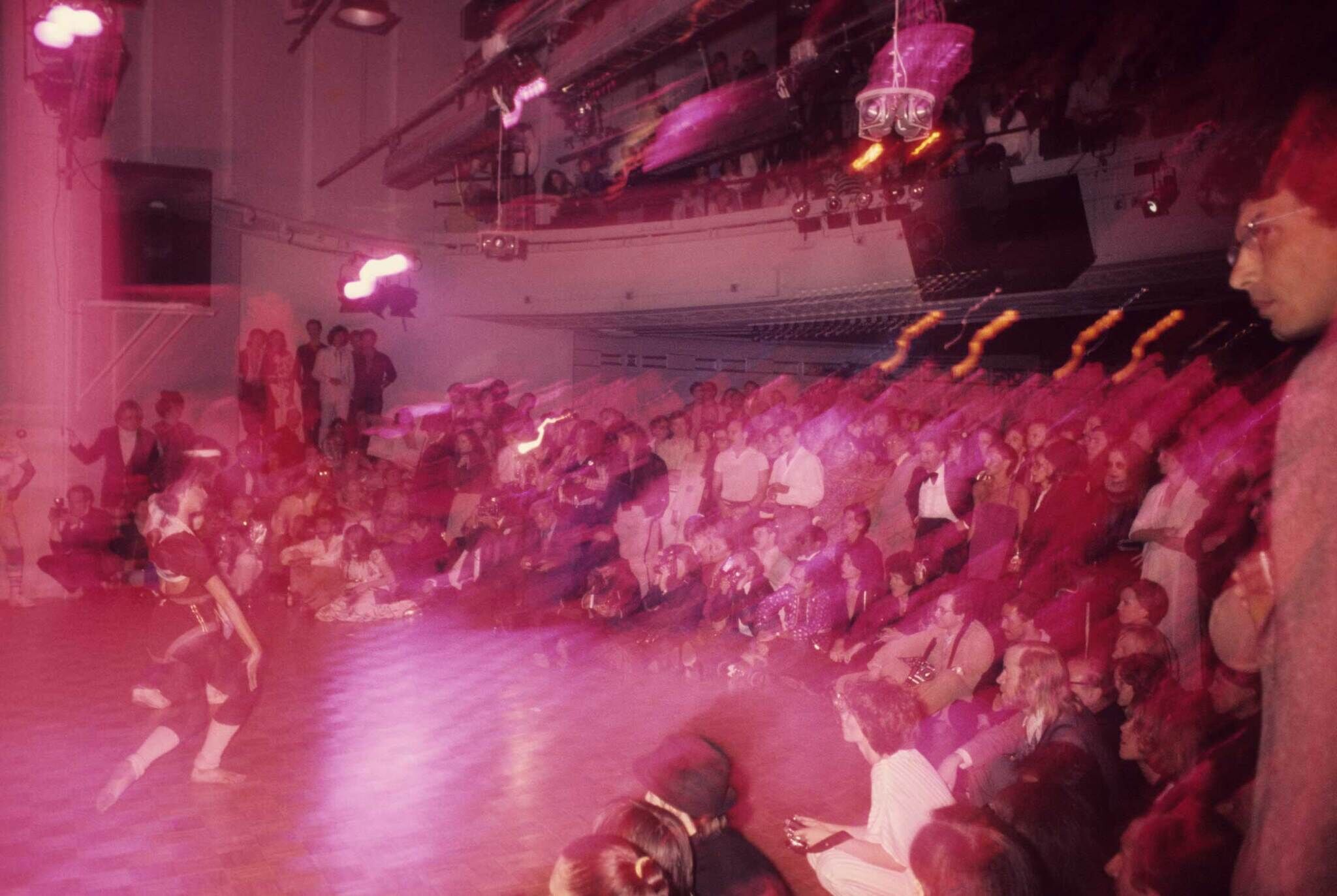 A crowded room with people watching a performance, bathed in pink and purple lighting, creating a blurred, dynamic effect.