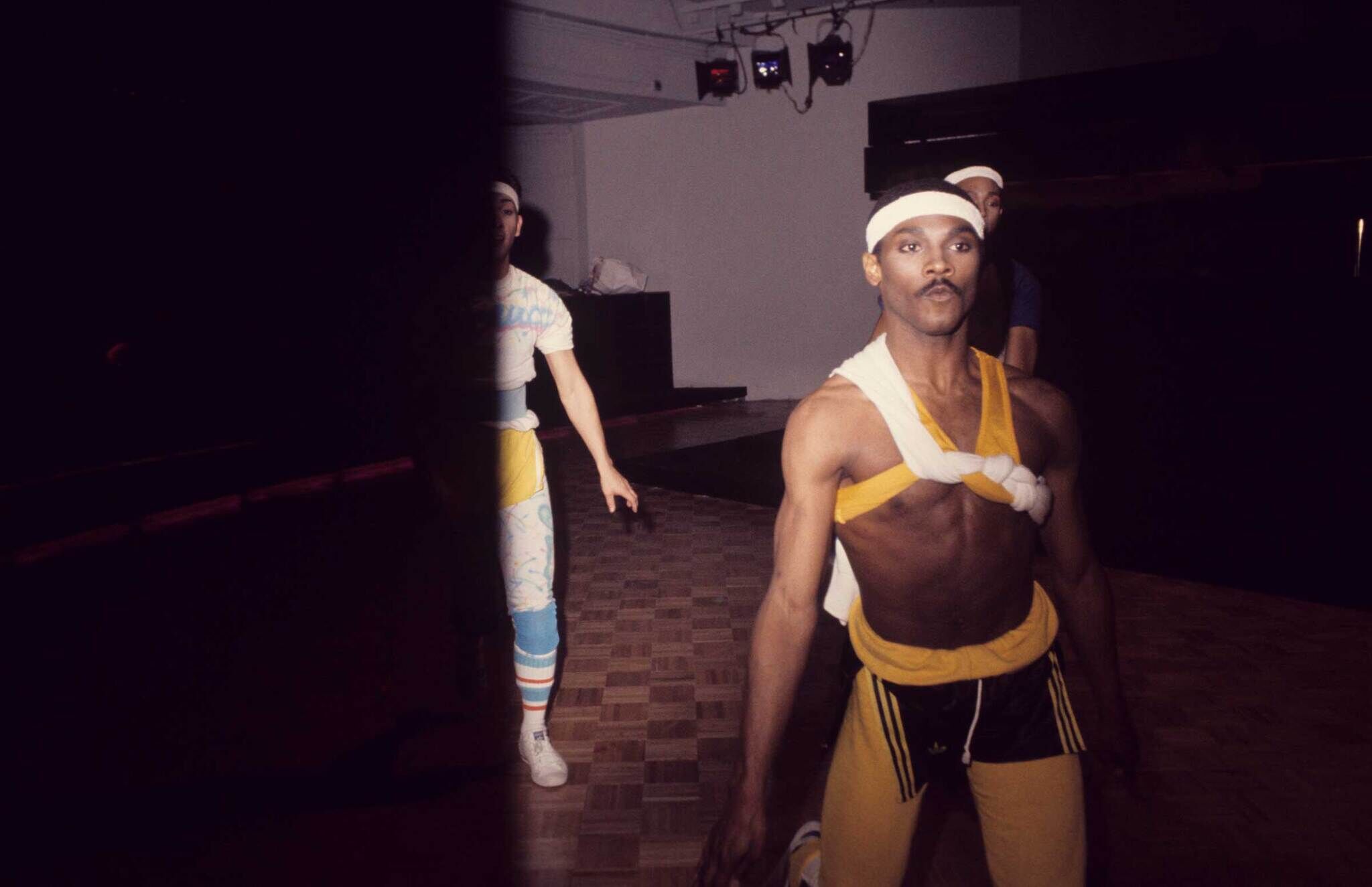 Three dancers in colorful athletic wear and headbands perform on a dimly lit stage, with one dancer prominently in the foreground.