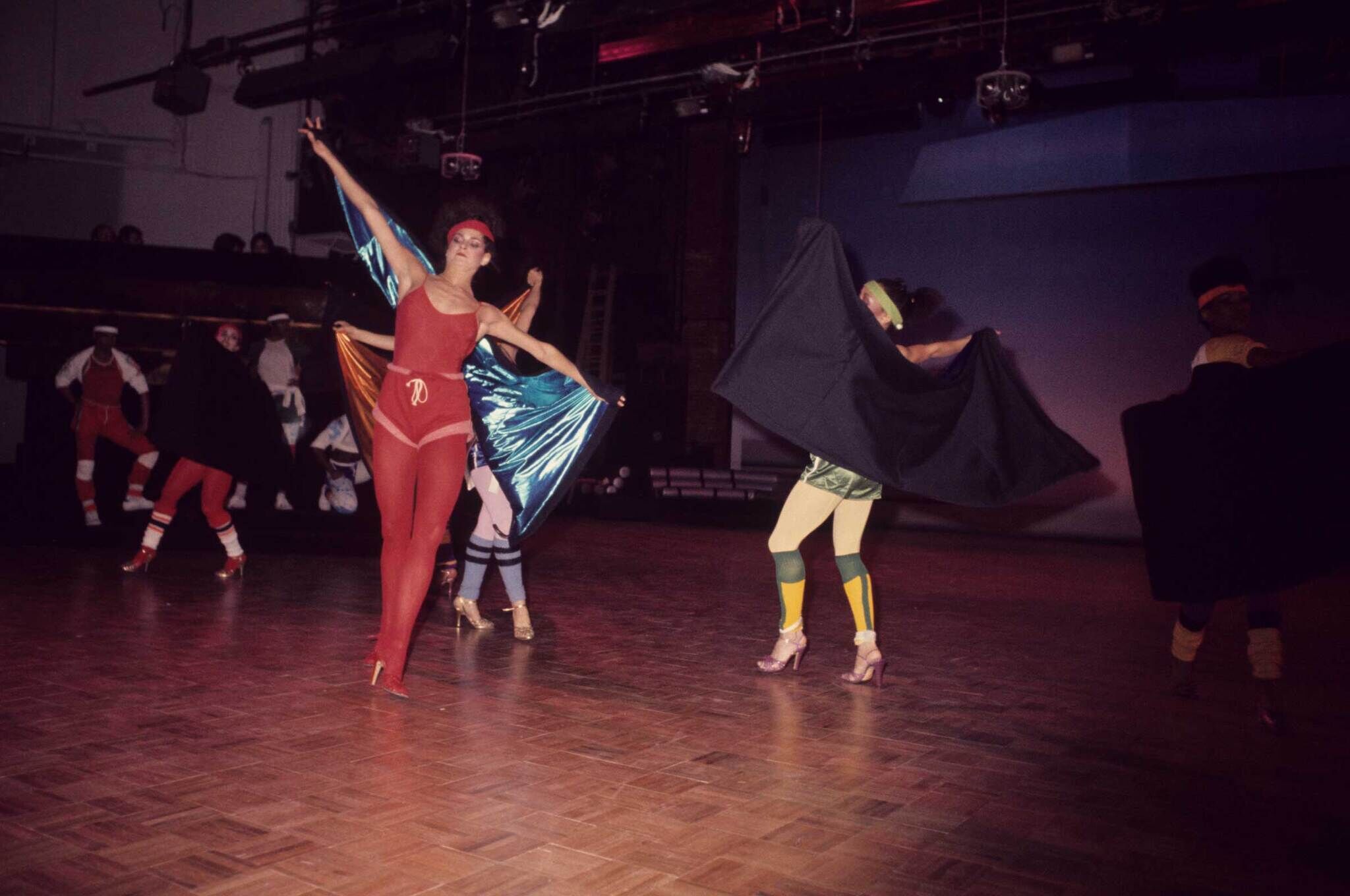 Dancers in colorful costumes perform on a dimly lit stage, with one dancer in red and others in various outfits, some holding capes.