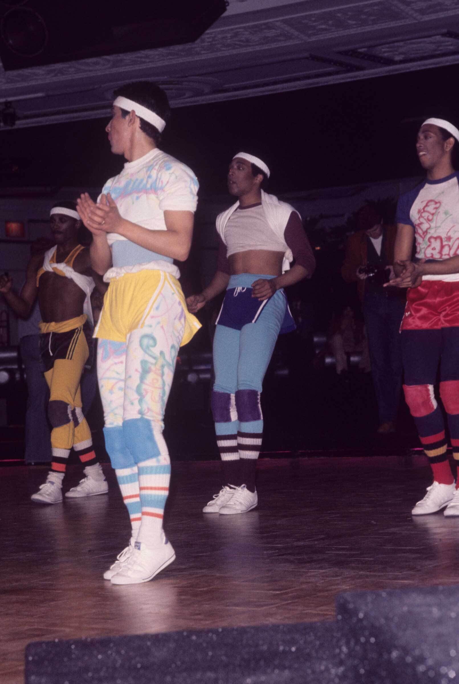 Group of people in colorful 80s workout attire, including headbands, leg warmers, and sneakers, dancing or exercising in a dimly lit room.