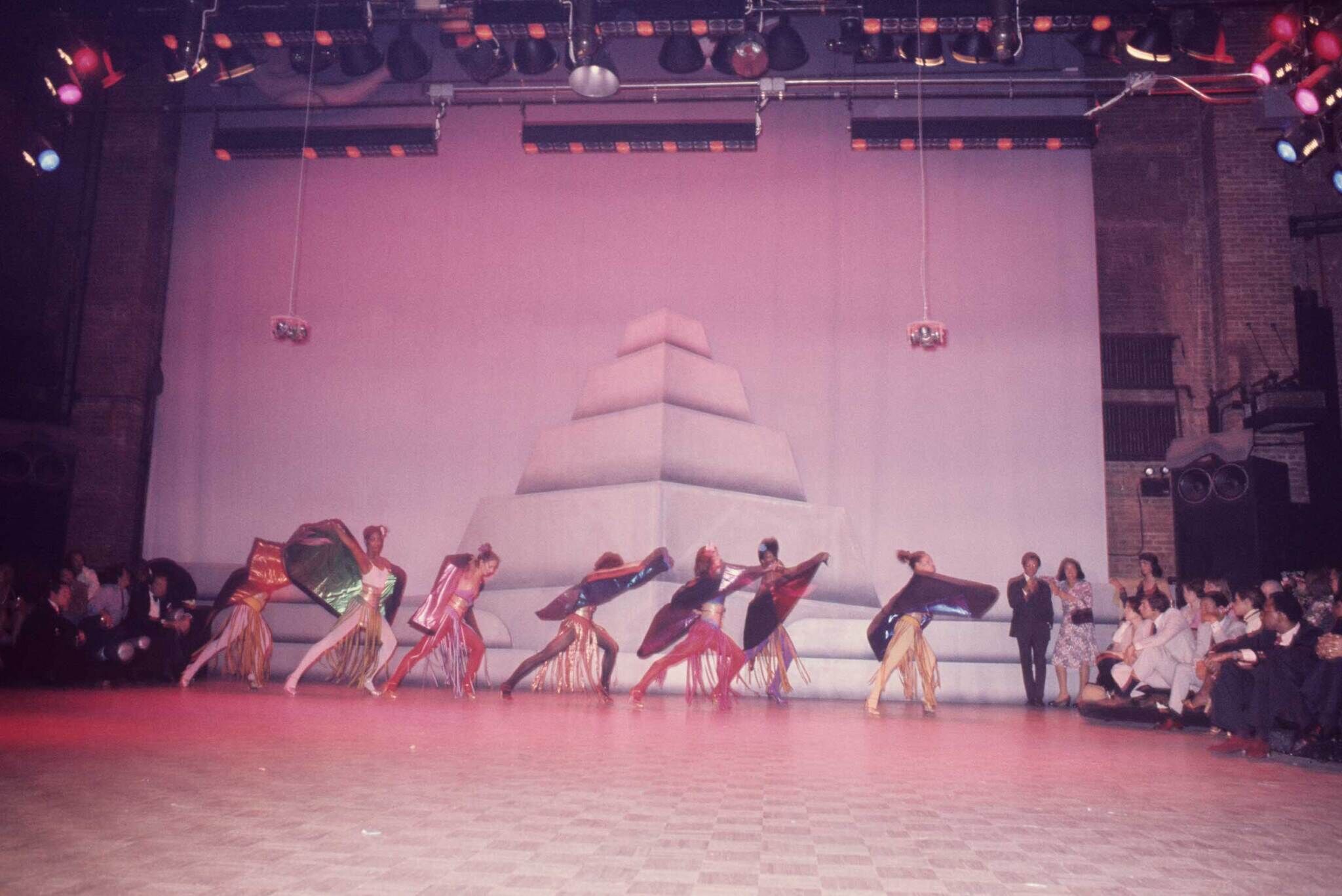 Dancers in colorful costumes perform on stage with a pyramid backdrop, while an audience watches from the sides.