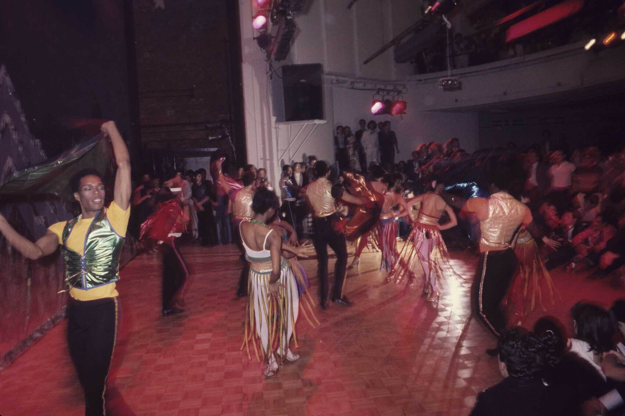 Dancers in colorful costumes perform energetically on a wooden floor, surrounded by a large audience in a dimly lit venue.