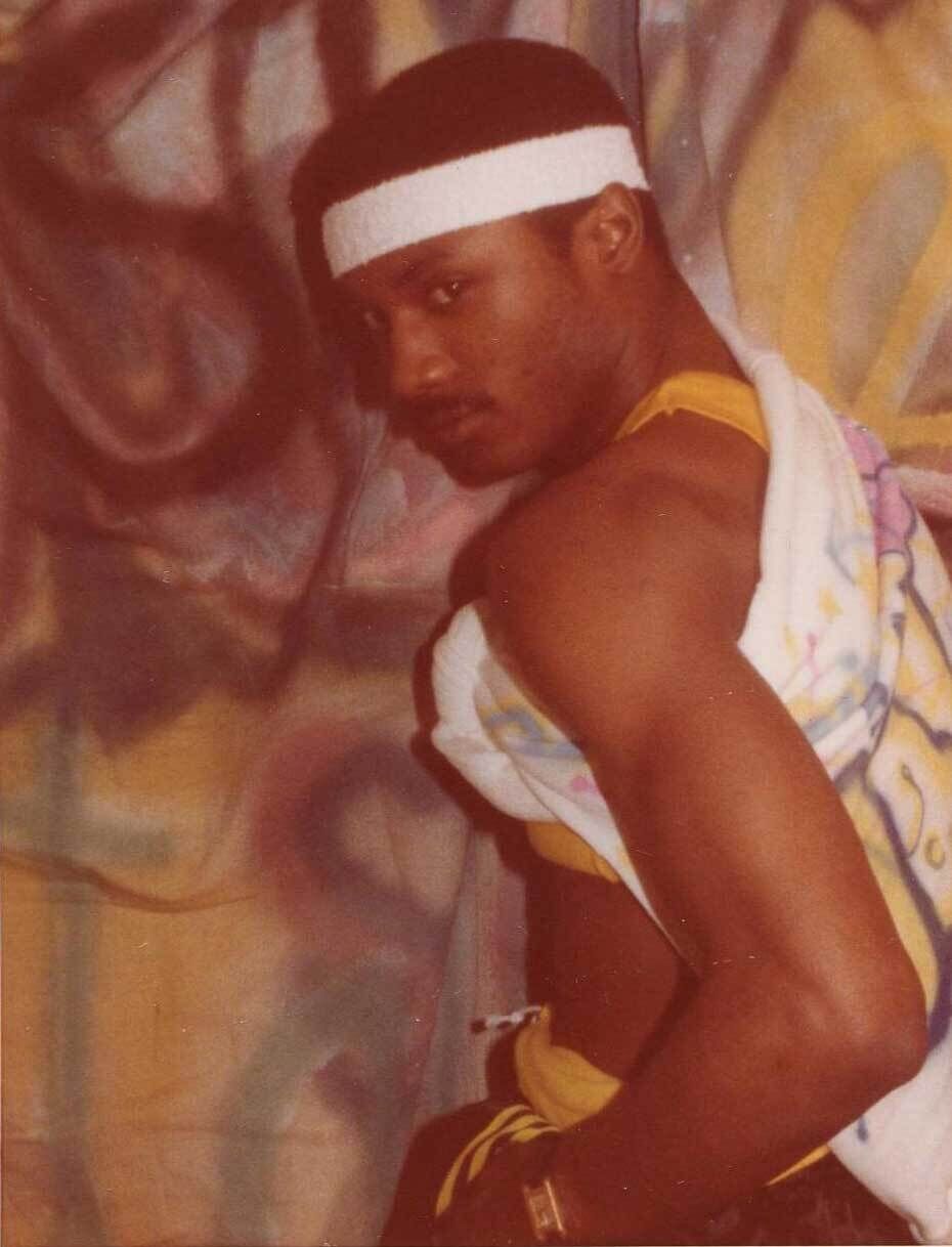 Man in a white headband and yellow tank top poses with a towel draped over his shoulder against a colorful, graffiti-style background.