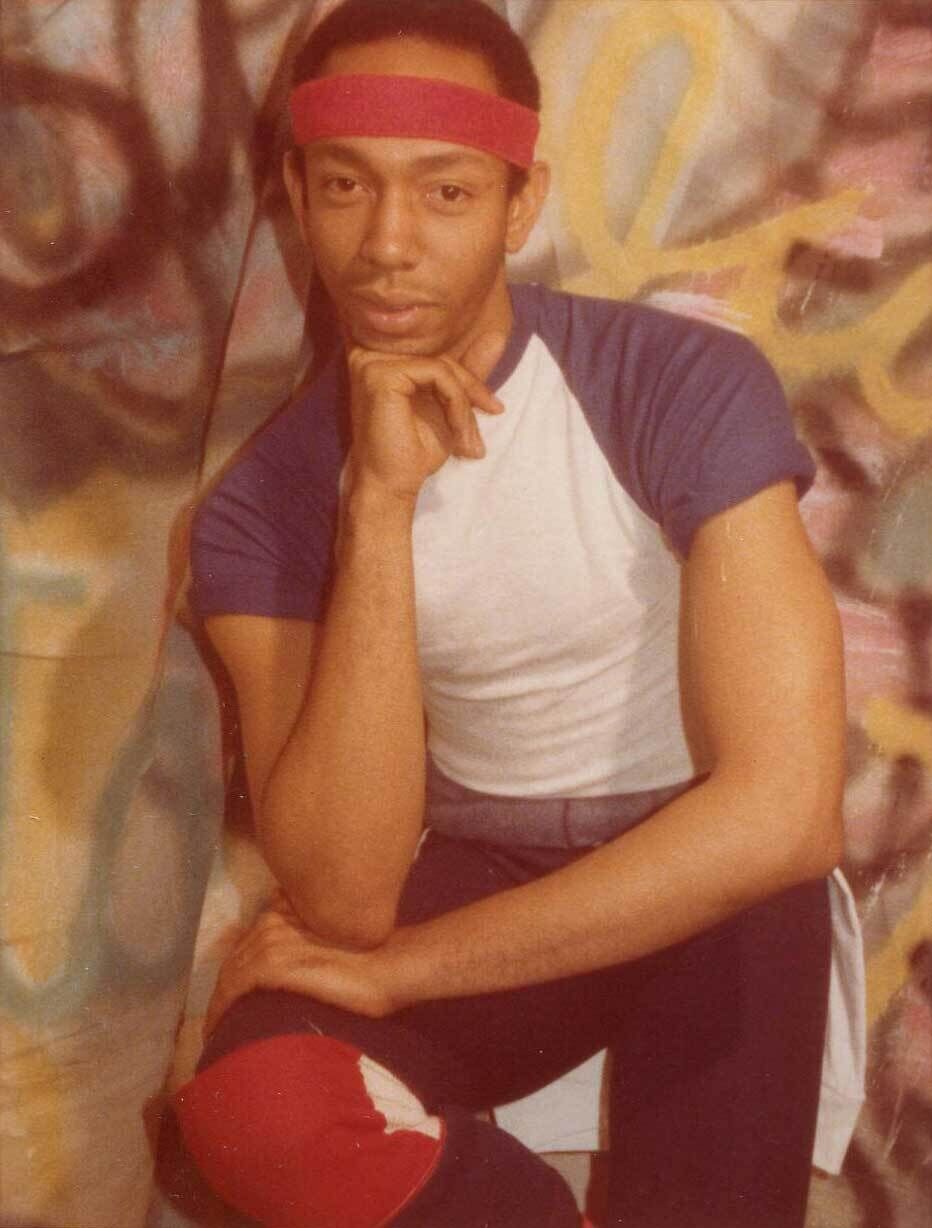 Person wearing a red headband and a white and blue shirt, posing with their chin resting on their hand against a graffiti-covered wall.