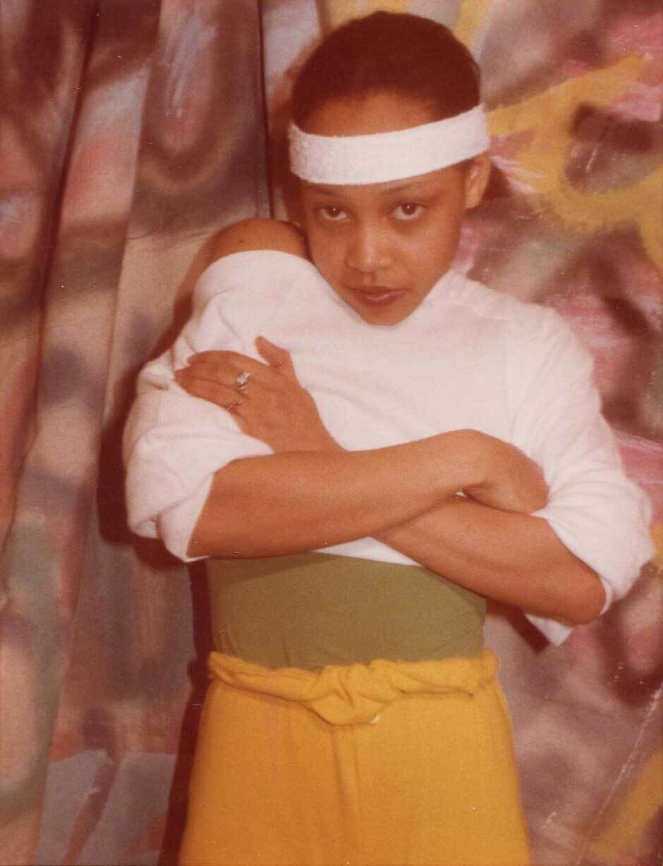 Person wearing a white headband, white top, and yellow pants, standing with arms crossed in front of a colorful, abstract background.